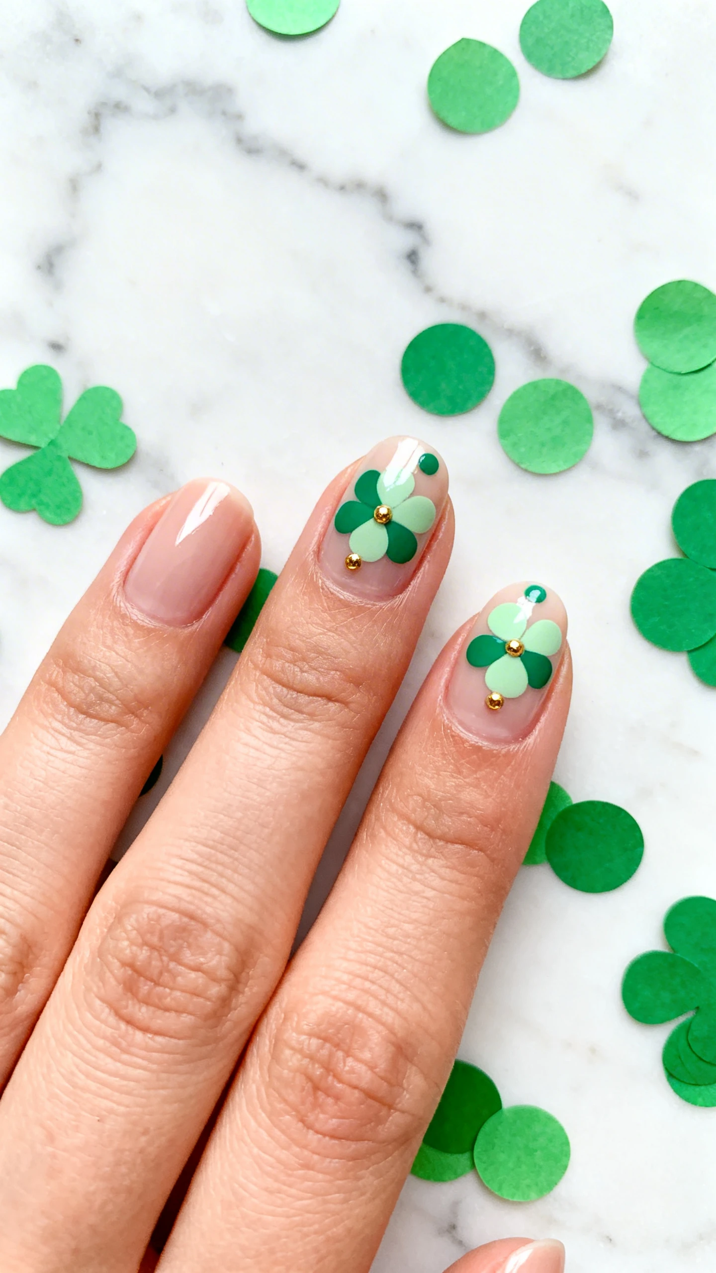5–1 mm, accent nails featuring tiny grouped dots in emerald and light sage arranged like a clover, optional single fine gold dot at each clover center, glossy topcoat; high-resolution, sharp focus. Background: minimalist marble slab with scattered confetti-like green paper circles for a festive Parisian touch; modern, Instagram-worthy; no face, only the hand and nails.