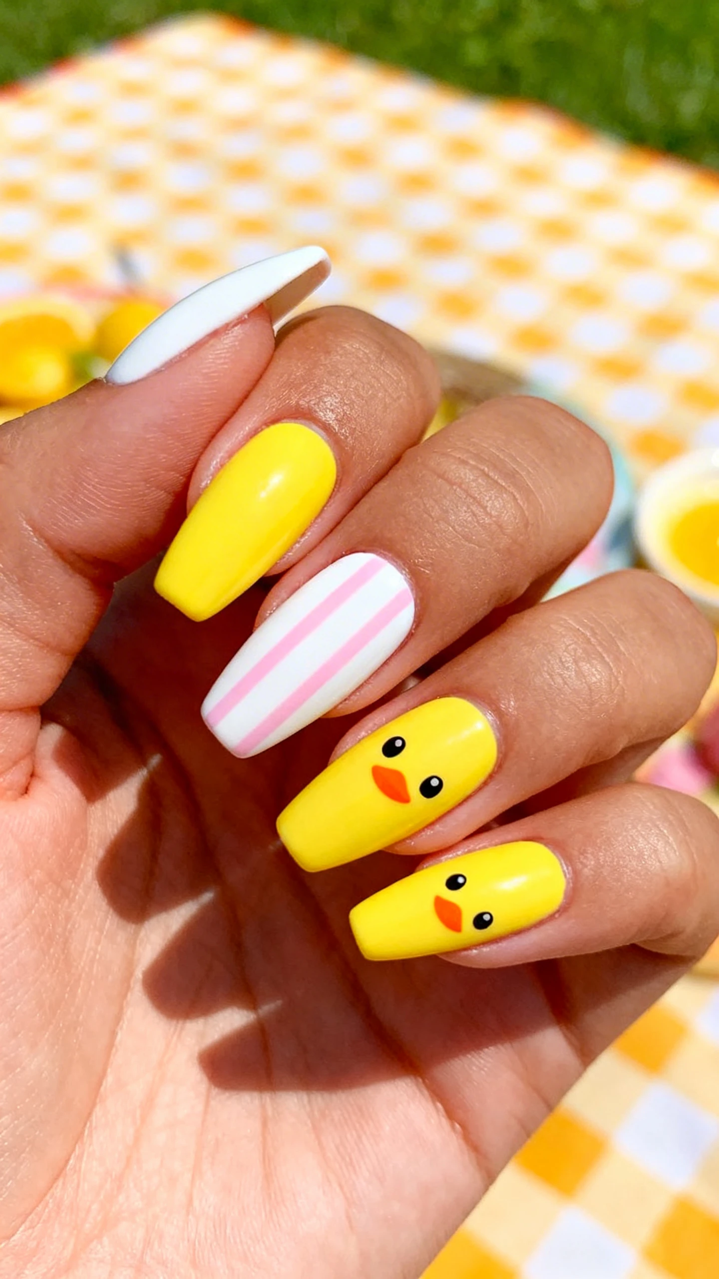 A beautiful macro photograph of a hand with alternating nails of bright sunshine yellow and crisp white. The white nails feature vertical pastel pink stripes, while the yellow nails feature cute chick faces with black outlines and orange beaks. High-resolution, sharp focus on the nails. Aesthetic background of a vibrant, sunny picnic setup with a gingham tablecloth. Modern, Instagram-worthy photography style. No faces visible, focus ONLY on the nails and hand., macro nail photography, high quality, Instagram-worthy, clean composition
