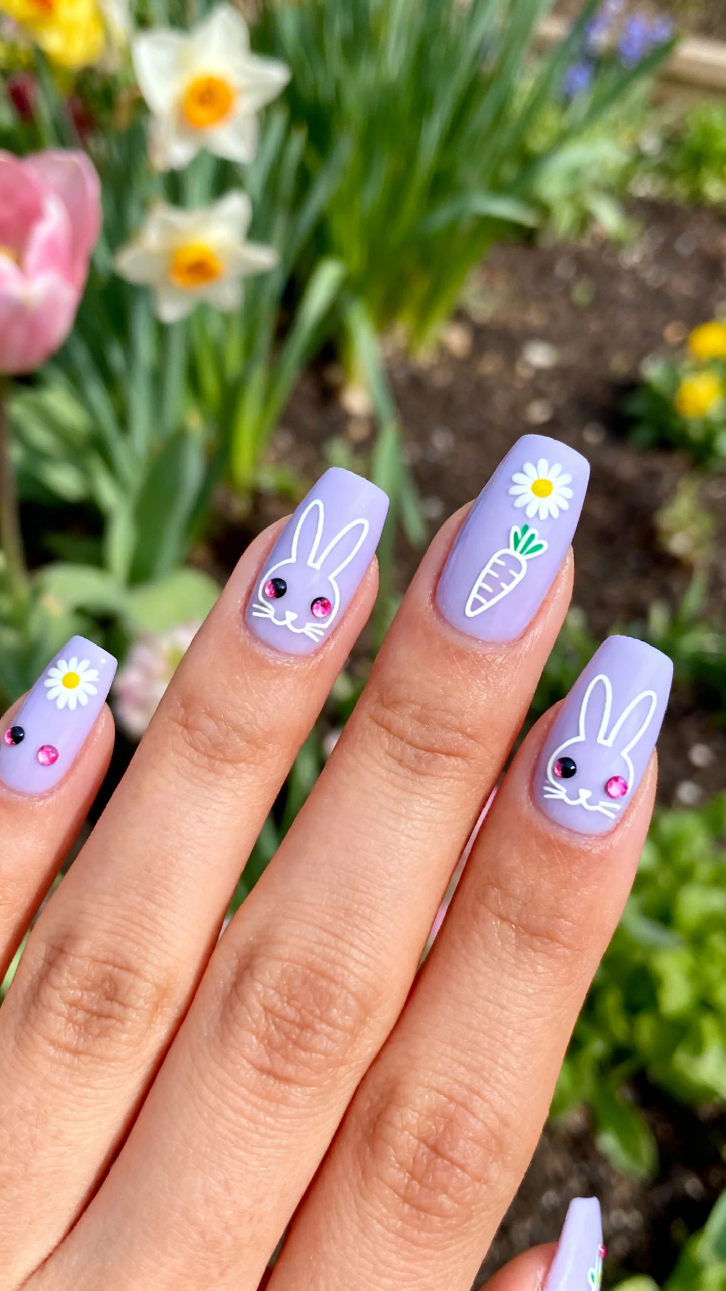 A beautiful macro photograph of a hand with nails coated in a soft, pastel lilac base. The nails display simple white line-art outlines of bunny faces and carrot silhouettes. Tiny round rhinestones in pink and black (0.5mm - 1mm) are used as the eyes for the bunnies, while others serve as the centers of tiny daisy flowers dotting the design. High-resolution, sharp focus on the nails. Aesthetic background featuring fresh spring flowers and garden greenery. Modern, Instagram-worthy photography style. No faces visible, focus ONLY on the nails and hand., macro nail photography, high quality, Instagram-worthy, clean composition