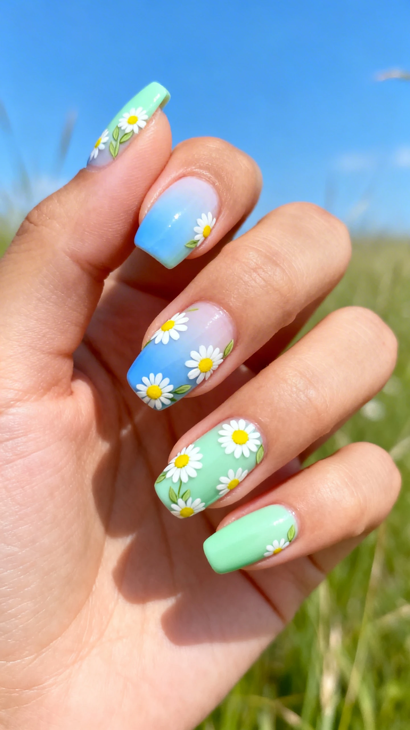 A beautiful macro photograph of a hand with nails featuring a smooth gradient blend from soft sky blue at the cuticle to mint green at the tip. Randomly scattered daisy clusters with white petals, bright yellow centers, and light green leaves adorn the nails near both the cuticle and tip. High-resolution, sharp focus on the nails. Aesthetic background of a bright, airy meadow with blue sky and soft focus grass. Modern, Instagram-worthy photography style. No faces visible, focus ONLY on the nails and hand., macro nail photography, high quality, Instagram-worthy, clean composition