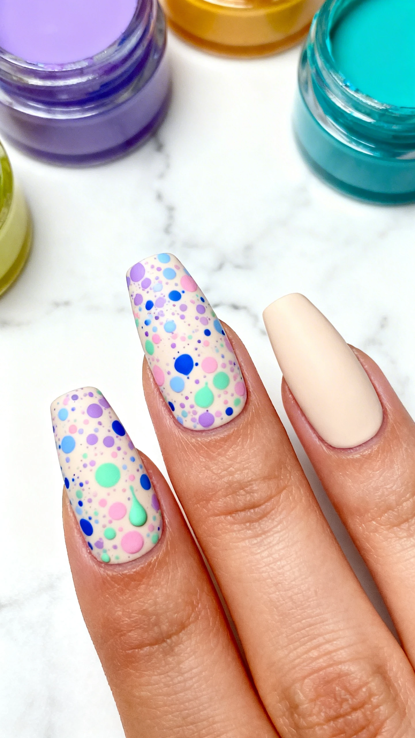 A beautiful macro photograph of a hand with smooth, neutral beige nails acting as an eggshell base. A dense, multidirectional splatter pattern in lavender, baby blue, soft pink, and mint green covers the nails, with splatters varying in size from tiny dots to larger drops. High-resolution, sharp focus on the nails. Aesthetic background of a clean, white marble surface with colorful paint jars. Modern, Instagram-worthy photography style. No faces visible, focus ONLY on the nails and hand., macro nail photography, high quality, Instagram-worthy, clean composition
