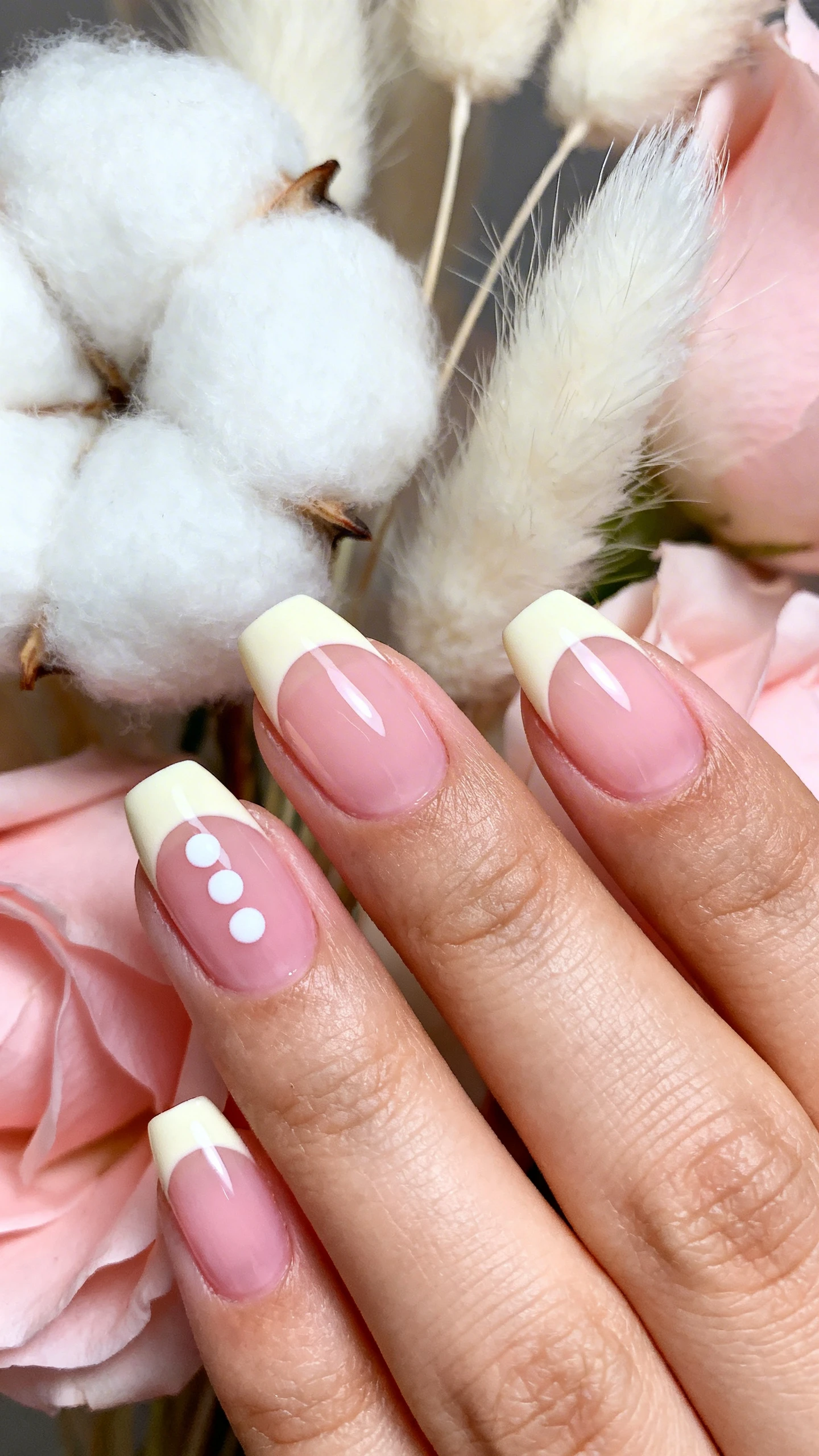 A beautiful macro photograph of a hand with soft, sheer baby pink jelly nails featuring ultra-thin creamy off-white French tips painted precisely at the very edge. The ring finger features three small white dots arranged in a cute cluster. High-resolution, sharp focus on the nails. Aesthetic background of fluffy white cotton tails and soft pastel pink petals. Modern, Instagram-worthy photography style. No faces visible, focus ONLY on the nails and hand., macro nail photography, high quality, Instagram-worthy, clean composition