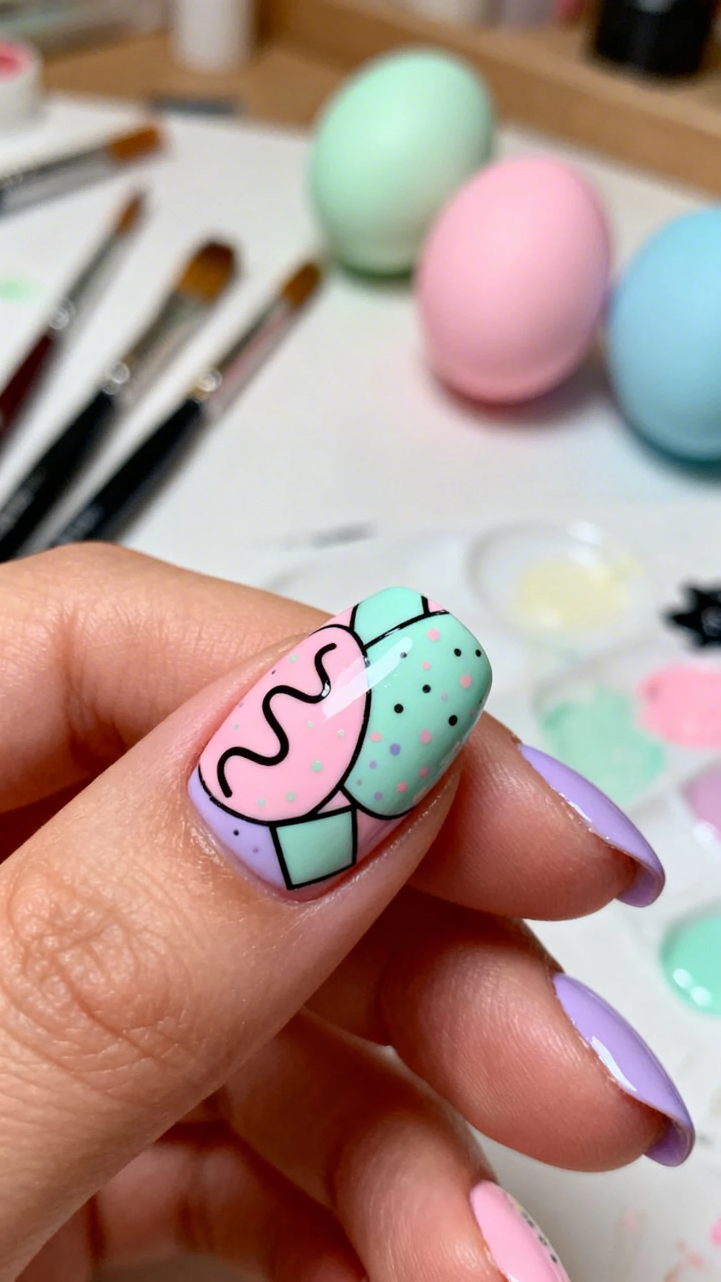 A beautiful macro photograph of one hand showing a mix and match nail design featuring abstract egg art with playful squiggles, geometric shapes, and speckled dots in pastel pink, mint green, and lavender gel polish. High-resolution, sharp focus on the nails. Aesthetic background of an artist's workspace with paintbrushes and colorful Easter eggs. Modern, Instagram-worthy photography style. No faces visible, focus ONLY on the nails and hand., macro nail photography, high quality, Instagram-worthy, clean composition