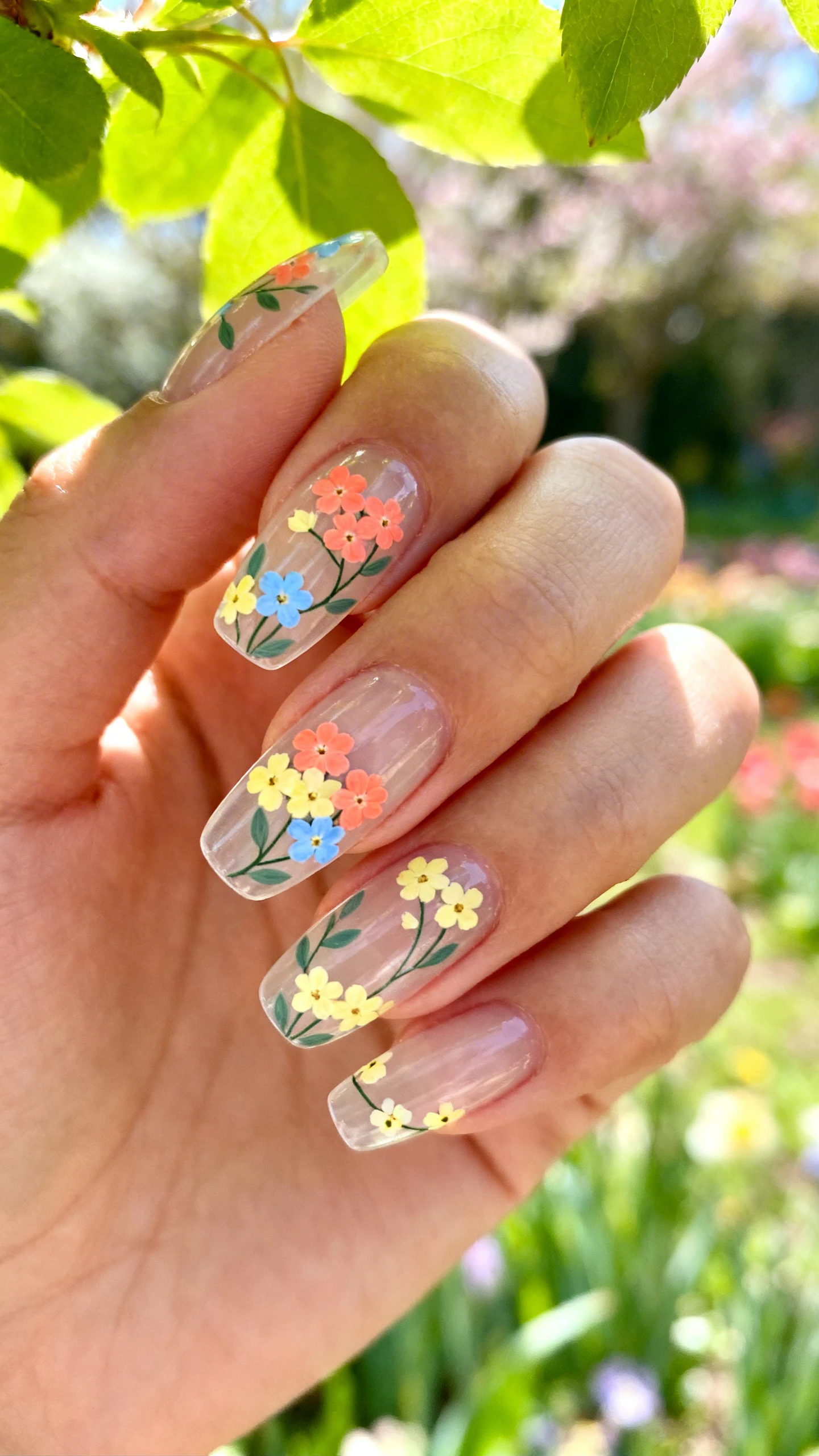 A beautiful macro photograph of one hand showing a set of press-on nails with a clear base. The nails feature tiny, detailed ditsy flowers painted in clusters, with five petals in coral, butter yellow, and sky blue, accented by sage green leaves and connecting vines. Some accent nails feature a full floral pattern, while others have minimalistic single blooms. The background is a lush, blurred spring garden with soft sunlight filtering through fresh green leaves. High-resolution, sharp focus on the nails. Modern, Instagram-worthy photography style. No faces visible, focus ONLY on the nails and hand., macro nail photography, high quality, Instagram-worthy, clean composition