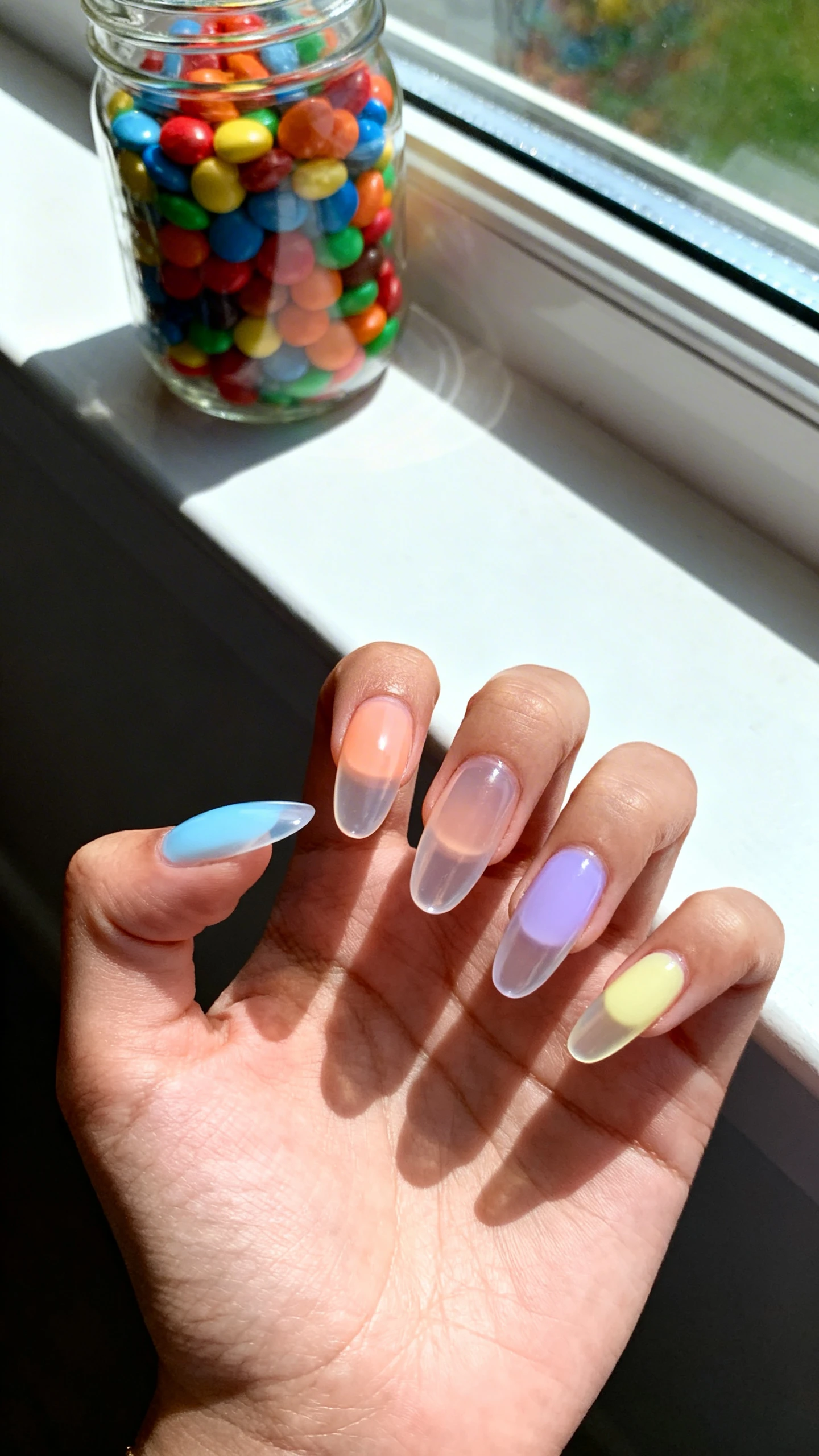 A beautiful macro photograph of one hand showing a sheer jelly skittles manicure where each nail features a different translucent pastel hue, including baby blue, soft peach, lilac, and pale yellow, with a high-gloss finish that catches the light. High-resolution, sharp focus on the nails. Aesthetic background of a bright, sunlit windowsill with a glass jar of colorful candies. Modern, Instagram-worthy photography style. No faces visible, focus ONLY on the nails and hand., macro nail photography, high quality, Instagram-worthy, clean composition