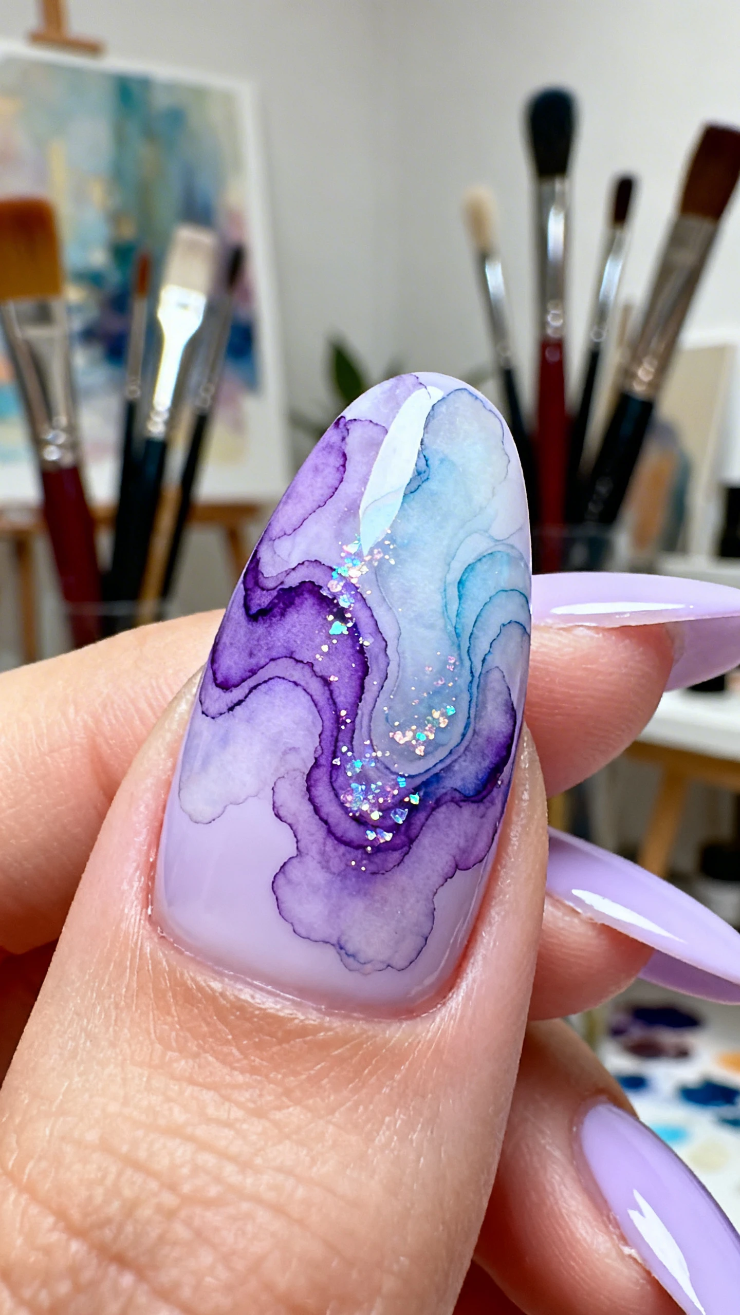 A beautiful macro photograph of one hand showing almond-shaped nails with a sheer, pale lilac cream base. Over the base, there are organic, fluid swirls in a deeper violet and a soft, periwinkle blue, resembling a translucent watercolor painting with fine iridescent glitter mixed into the swirls. High-resolution, sharp focus on the nails. Aesthetic background of a modern art studio with soft purple lighting and paintbrushes. Modern, Instagram-worthy photography style. No faces visible, focus ONLY on the nails and hand., macro nail photography, high quality, Instagram-worthy, clean composition