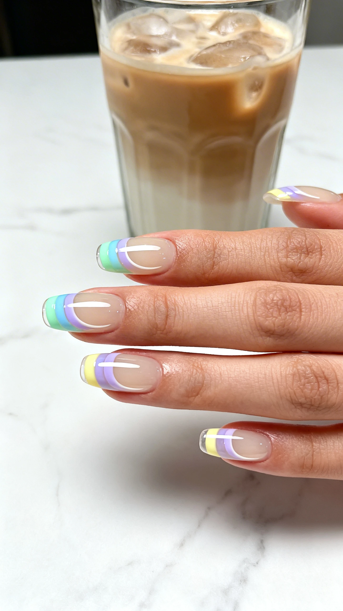 A beautiful macro photograph of one hand showing a set of press-on nails with a translucent, milky beige jelly base. The tips feature a thick, opaque stripe in pastel colors including mint green, baby blue, soft lavender, and pale yellow, with a crisp and curved smile line. The finish is a shiny, glass-like gel. The background is a minimalist, sleek white marble surface next to a glass of iced latte. High-resolution, sharp focus on the nails. Modern, Instagram-worthy photography style. No faces visible, focus ONLY on the nails and hand., macro nail photography, high quality, Instagram-worthy, clean composition