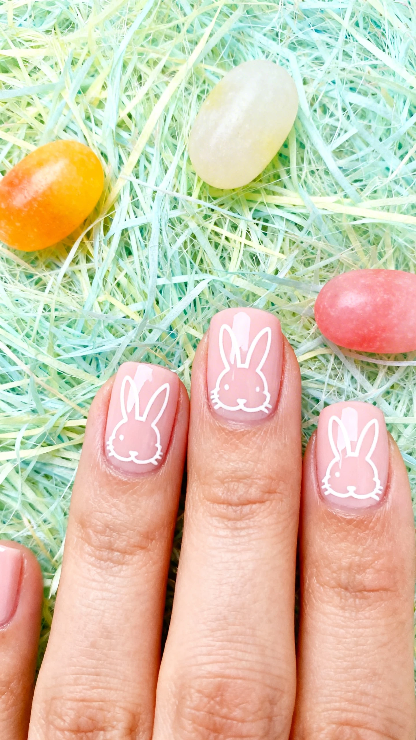 A beautiful macro photograph of one hand showing a minimalist bunny nail design featuring simple white line art bunny silhouettes on a glossy pale pink base in detail. High-resolution, sharp focus on the nails. Aesthetic background featuring pastel Easter grass and a few colorful jellybeans. Modern, Instagram-worthy photography style. No faces visible, focus ONLY on the nails and hand., macro nail photography, high quality, Instagram-worthy, clean composition