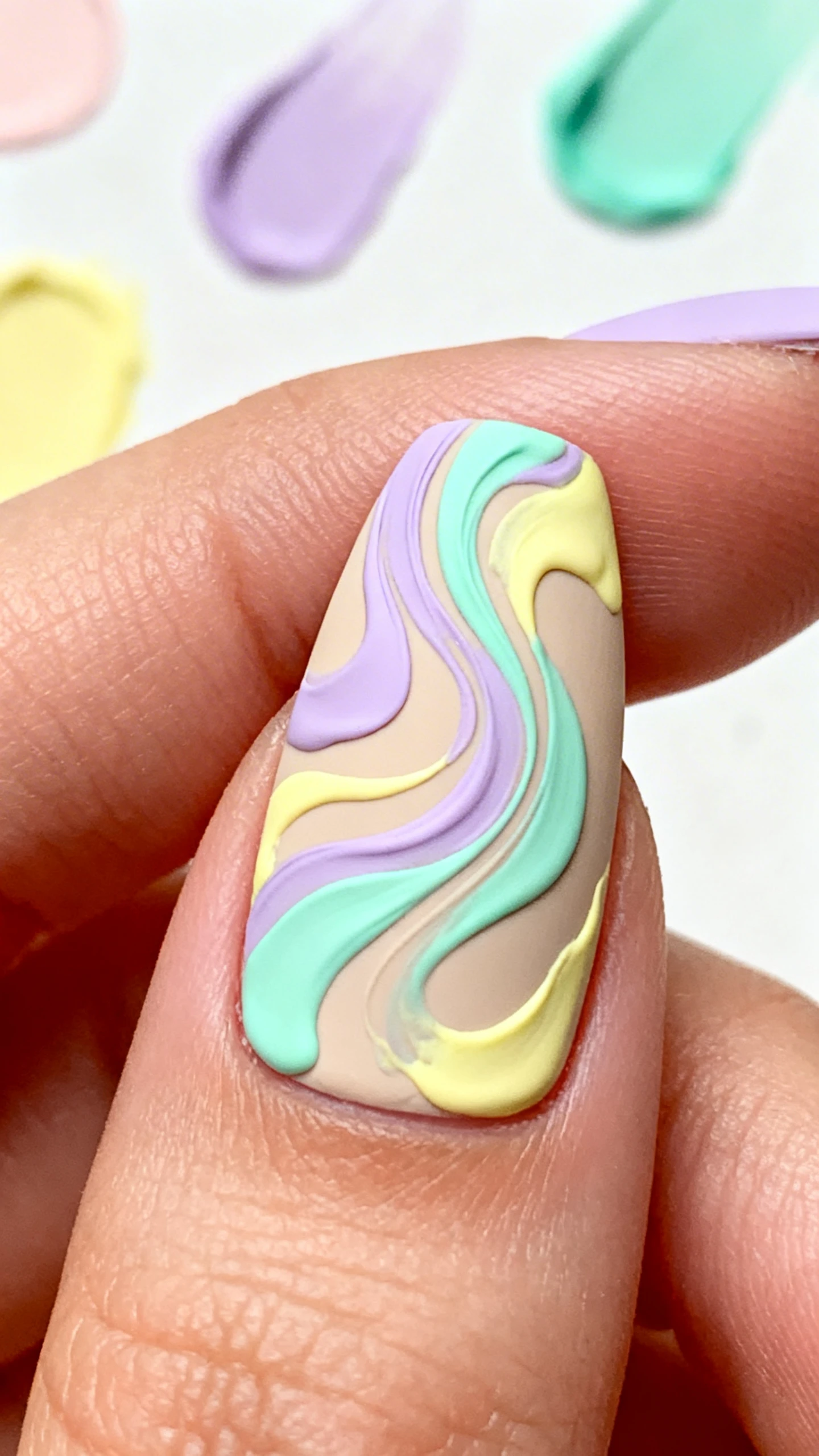 A beautiful macro photograph of one hand showing an abstract swirl nail design. Thick, fluid brushstrokes of pastel lilac, mint green, and butter yellow colors mix slightly at the edges as they swirl across a neutral beige base. The entire nail is covered in a matte top coat. High-resolution, sharp focus on the nails. Aesthetic background of pastel art supplies or abstract colorful shapes. Modern, Instagram-worthy photography style. No faces visible, focus ONLY on the nails and hand., macro nail photography, high quality, Instagram-worthy, clean composition