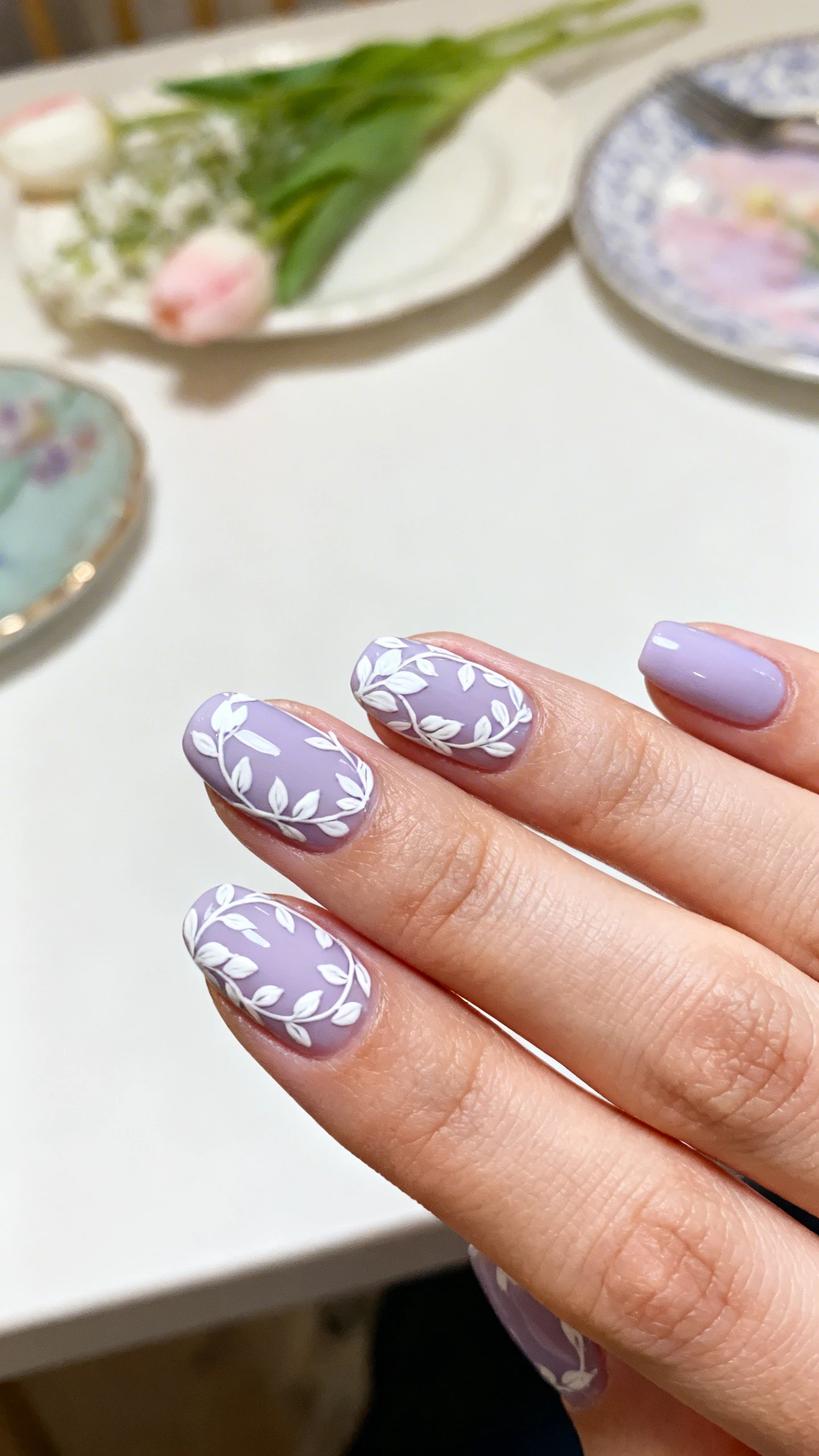 A beautiful macro photograph of one hand showing a cohesive spring manicure featuring a soft lilac base with intricate hand-painted white floral vines and leaves wrapping around the cuticles, representing the advanced artistry achievable with quality brushes. The background is a minimalist, elegant spring table scape. High-resolution, sharp focus on the nails, modern Instagram-worthy photography style, no faces visible, focus ONLY on the nails and hand., macro nail photography, high quality, Instagram-worthy, clean composition