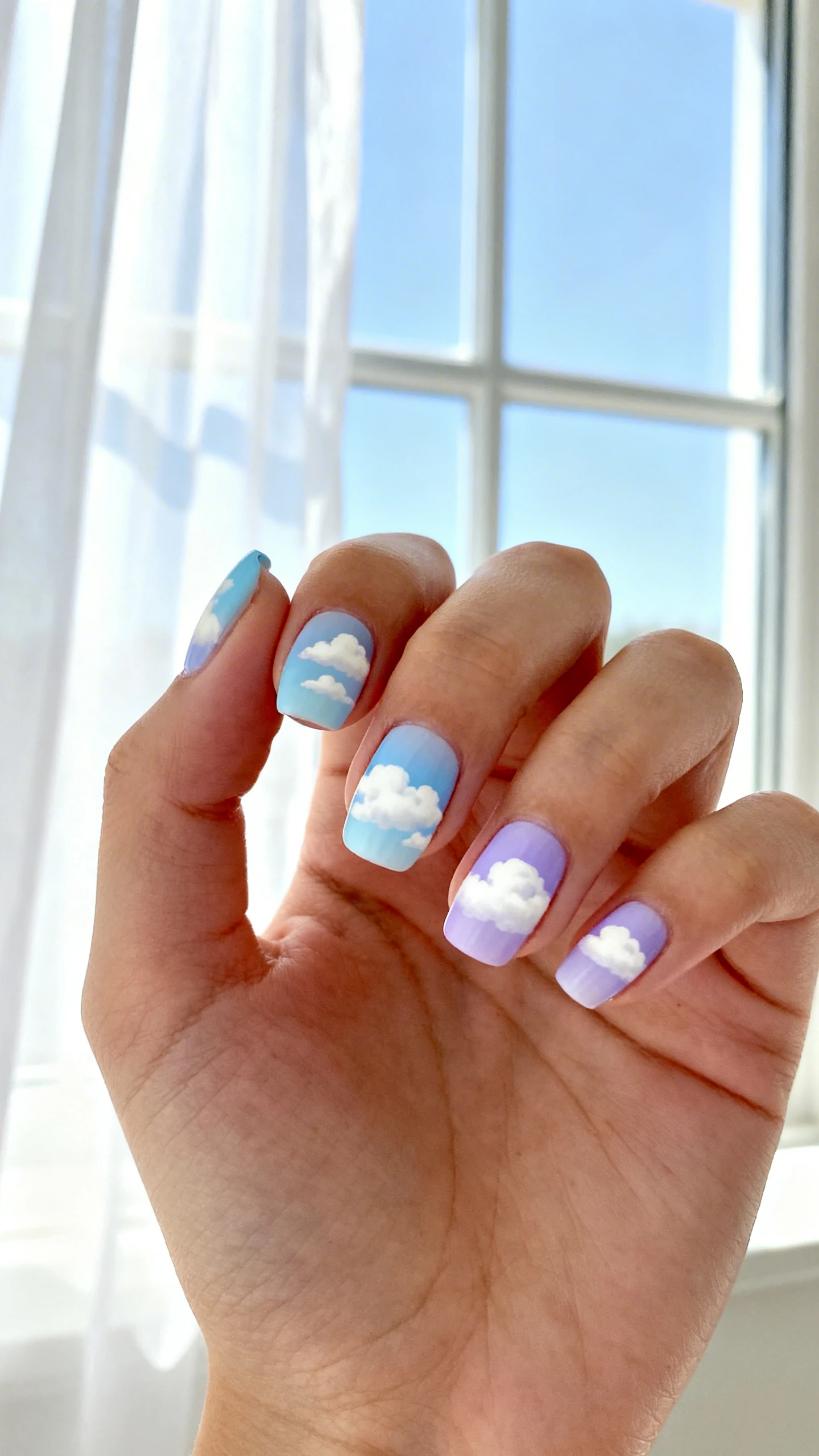 A beautiful macro photograph of one hand showing dreamy pastel sky nail art. The nails are painted with a soft gradient of baby blue and lavender, featuring fluffy white hand-painted clouds floating across the tips. High-resolution, sharp focus on the nails. The background is a bright, airy window view with sheer white curtains. Modern, Instagram-worthy photography style. No faces visible, focus ONLY on the nails and hand., macro nail photography, high quality, Instagram-worthy, clean composition