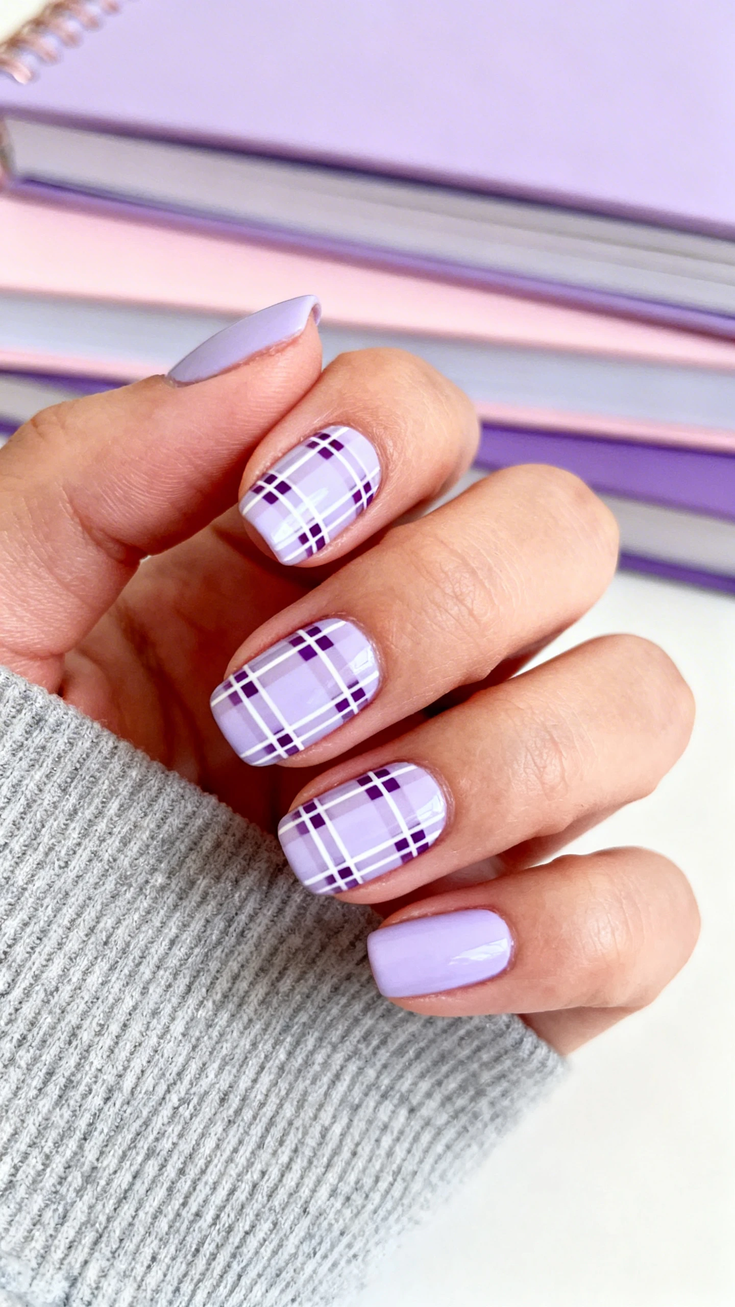 A beautiful macro photograph of one hand showing preppy pastel plaid nail art. The nails are painted in a soft lavender base with a classic crisscross plaid pattern in thin white and deeper purple stripes. High-resolution, sharp focus on the nails. The background is a cozy, blurred stack of pastel and lavender notebooks. Modern, Instagram-worthy photography style. No faces visible, focus ONLY on the nails and hand., macro nail photography, high quality, Instagram-worthy, clean composition