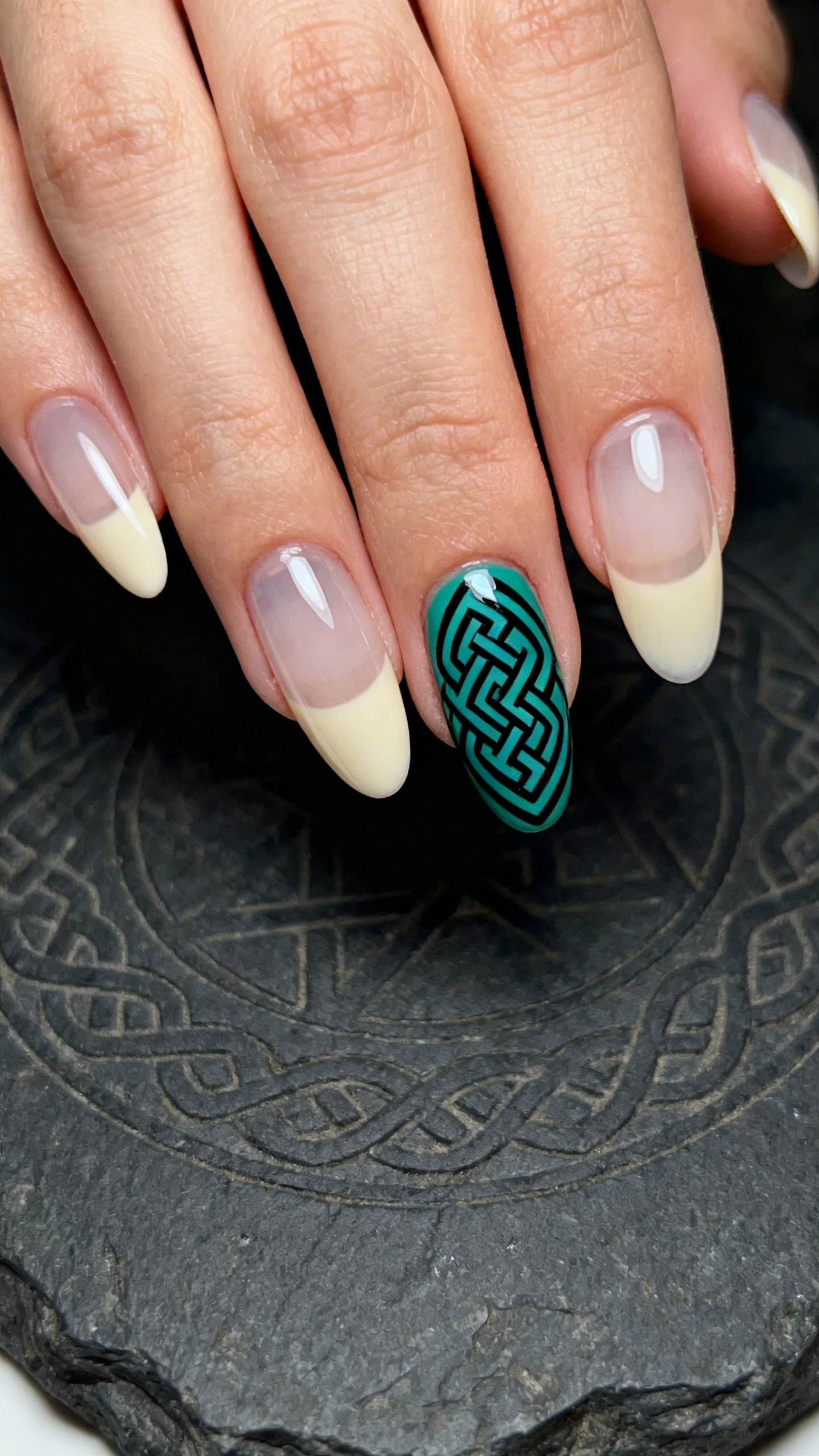 A beautiful macro photograph of one hand with medium almond nails featuring mostly clear/lightly tinted cream bases topped glossy; a single accent nail (ring finger) displays precise interlaced Celtic knotwork in deep emerald and black, with thin, even lines and intentional transparent negative spaces; ultra-clean edges and symmetry; high-resolution, sharp focus; aesthetic background: dark stone slab with a faint Celtic pattern engraving softly out of focus; moody, modern lighting; no faces, only the hand and nails.