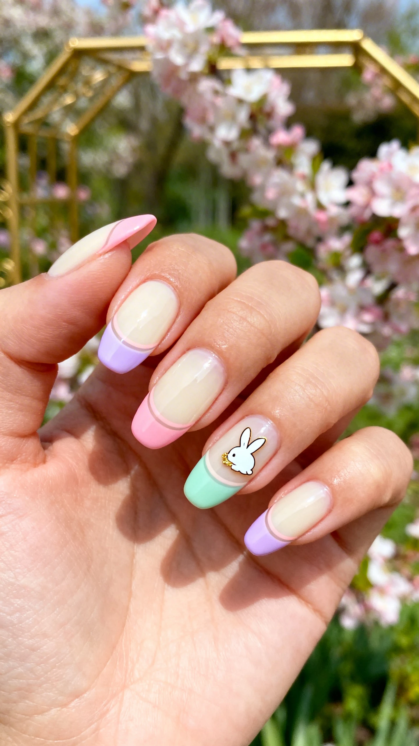 A beautiful macro photograph of one hand with medium almond nails in a glassy glossy finish, sheer cream base on all nails. Ultra-thin pastel micro-French tips in blush pink, lavender, and mint, alternating across nails with perfectly curved, delicate lines; a single small gold-foil-accented white bunny decal near the sidewall of the ring finger for a “gilded garden” touch. High-resolution, crisp focus to highlight the fine French lines and subtle gold gleam; softly blurred garden background with pastel blossoms and a hint of gilded frame or garden trellis. Clean, modern Instagram lighting; no faces, only the hand and nails.