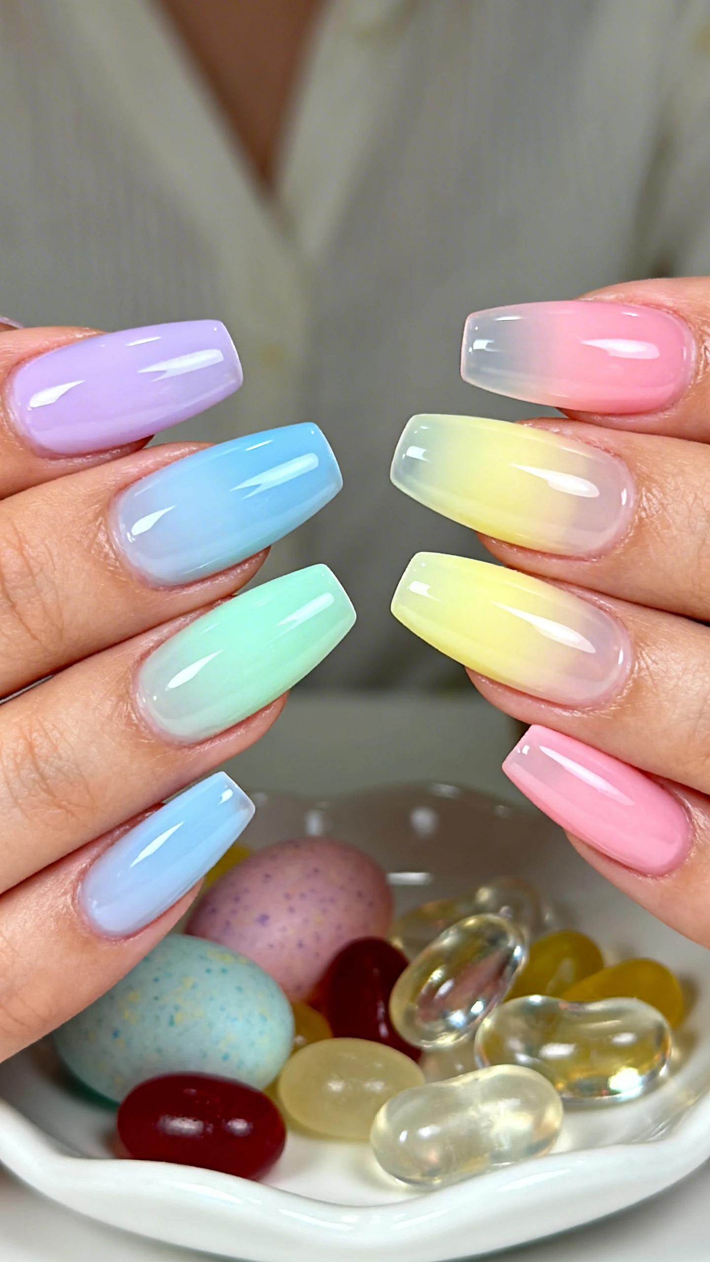 A beautiful macro photograph of one hand with short to medium almond press-on nails in a high-gloss gel finish, each nail a sheer jelly ombré fading from cuticle to tip; left hand arranged as soft lilac, powder blue, mint green, lemon sorbet, and blush pink, mirrored on the right. Capture the smooth gradient transitions and translucent jelly depth in sharp focus. Background: softly blurred pastel Easter candy and glass jelly beans on a white ceramic dish for a dreamy, spring vibe. Modern, Instagram-worthy lighting, no faces, only the hand and nails centered.