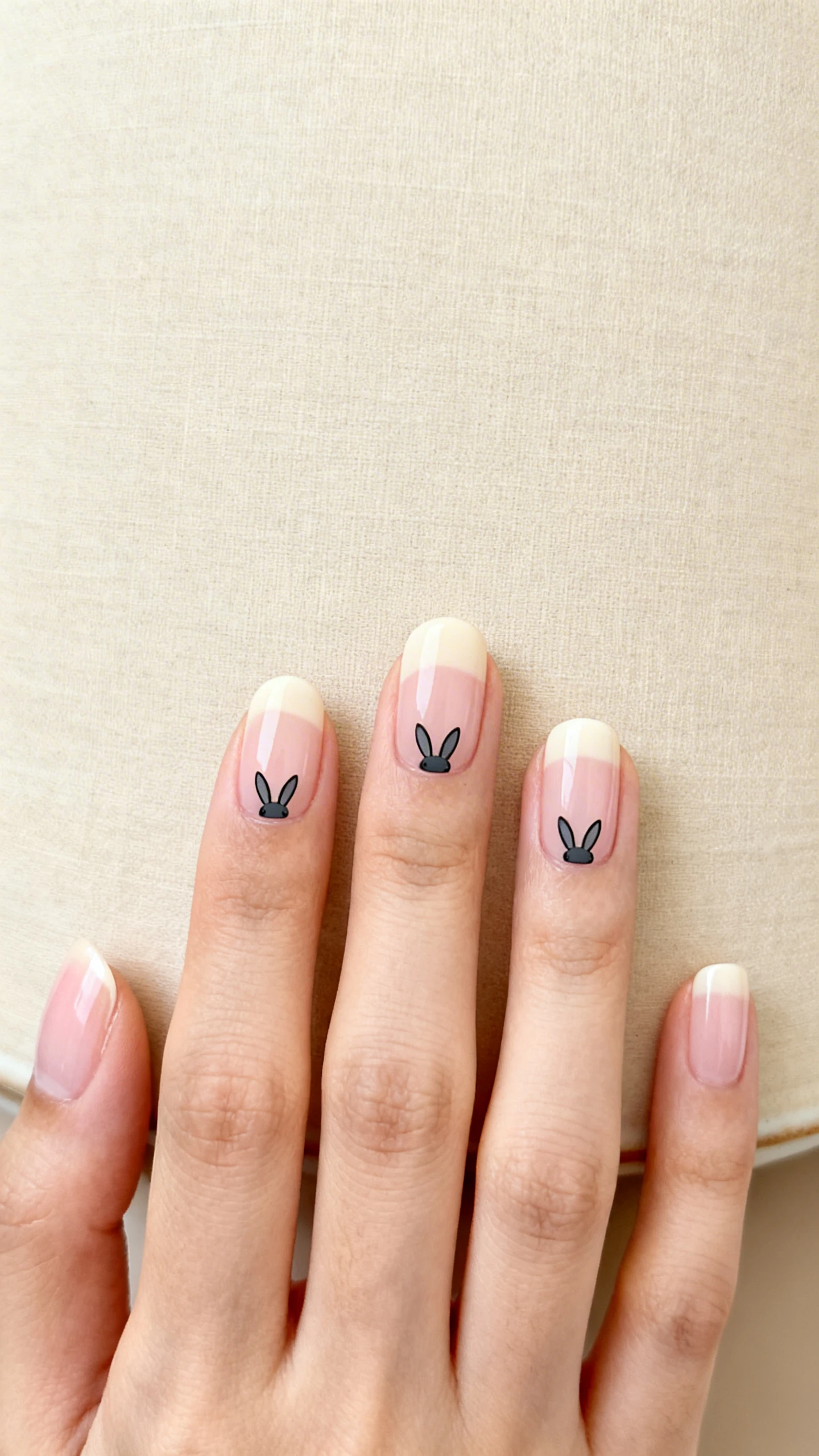 A beautiful macro photograph of one hand with short to medium round nails, thin even free edge, all painted in a sheer cream/milky pink base for a translucent wash. Minimalist tiny black or charcoal-gray bunny ear decals placed at the cuticle line on the index and ring fingers, ears angled slightly inward as if peeking over the cuticle. High-resolution, sharp focus on the subtleties of the sheer polish and clean cuticles; minimalist background of soft beige linen or matte ceramic for a calm, modern vibe. Instagram-worthy studio lighting; no faces, only the hand and nails.