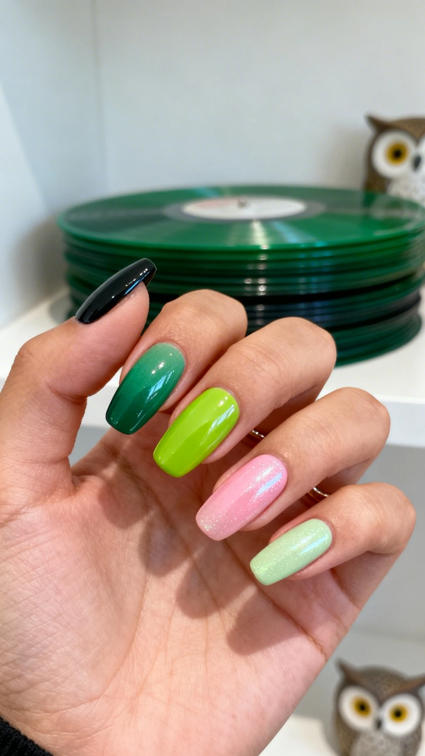 A beautiful macro photograph of one hand with short-to-medium squoval nails in sharp focus, showing a Skittles gradient across five fingers. Palette arranged darkest thumb to lightest pinky: thumb in blackened pine green cream, index in emerald green cream, middle in true Kelly green cream, ring in vibrant lime green cream, pinky in pale pistachio-mint cream. Optional ultra-thin glossy top coat for uniform shine and a subtle pearly glazed topper to tie the set together. High-resolution, no faces visible. Background: minimalist studio with stacked green vinyl records and a subtle OVO-owl hint in decor, Instagram-worthy lighting.