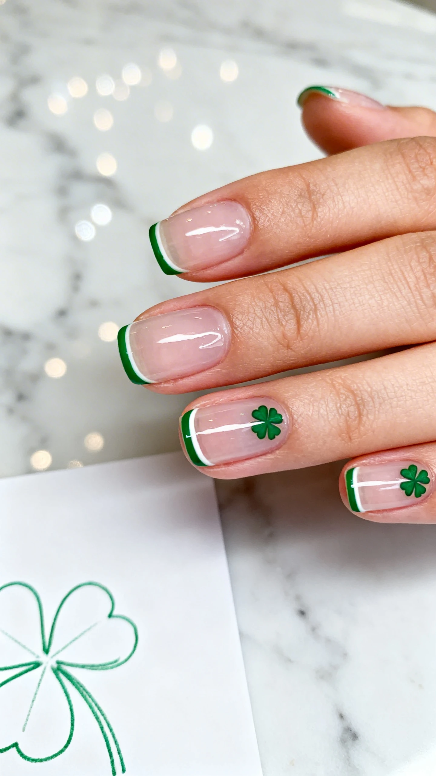 A beautiful macro photograph of one hand with short square or short almond nails; translucent pinkish-clear base for a healthy gloss; each nail has a thin shamrock-green French outline tracing just the free edge; ring and index fingers feature two micro four-leaf clover half-moons nestled along the cuticle line; high-resolution, sharp focus to show the precise outline and micro clovers; background is a clean marble surface with a delicate green line-art shamrock sketch pad partially visible in bokeh; modern minimalist composition, Instagram-ready, no faces, only the hand.