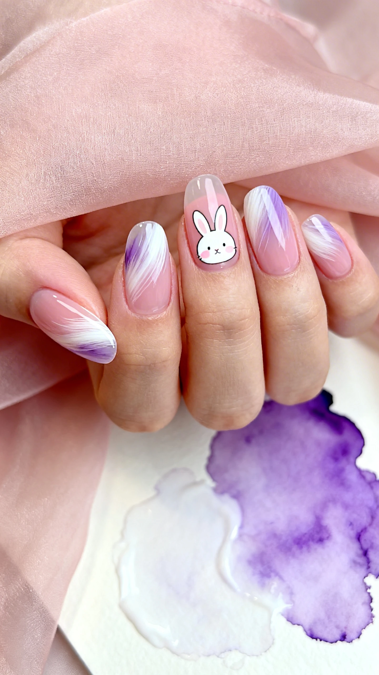 A close-up macro photograph of one hand with short almond press-ons on a sheer blush-pink base. Most nails feature semi-transparent, feathery brushstrokes in pastel lilac and white sweeping diagonally from lower sidewall to opposite tip, softly diffused for painterly motion. One accent nail shows a minimal, hand-painted white bunny silhouette with a tiny blush nose and subtle ear shading, small and off-center for elegance. Emphasize the translucent layers and soft edges. Background: blush chiffon and a watercolor palette with lilac/white puddles, softly out of focus. Clean, Instagram-ready style; no faces.
