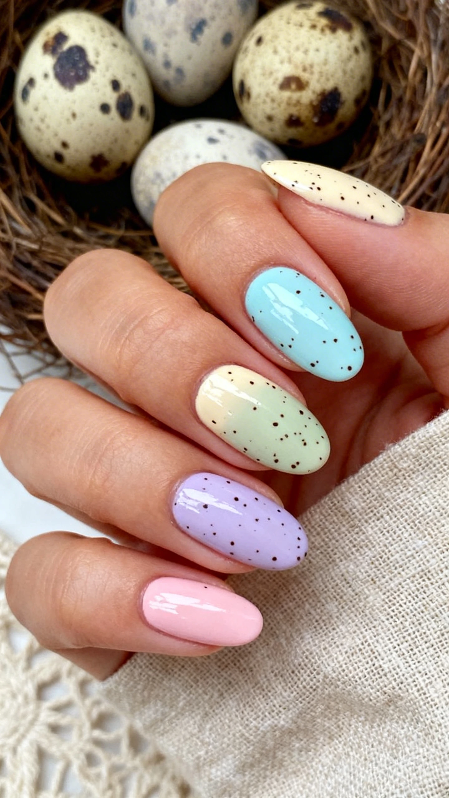 A crisp macro shot of one hand with medium oval nails featuring soft pastel creme bases in pale robin’s egg blue, light cream, muted sage, lilac, and blush pink; each nail has fine black and dark brown micro-speckles scattered randomly with slightly varied density nail to nail; sealed with a high-shine top coat for subtle shimmer; tight, high-resolution focus on the speckling details; softly blurred background of speckled eggs in a nest and linen textures; bright, airy editorial lighting; no faces, only the hand and nails.