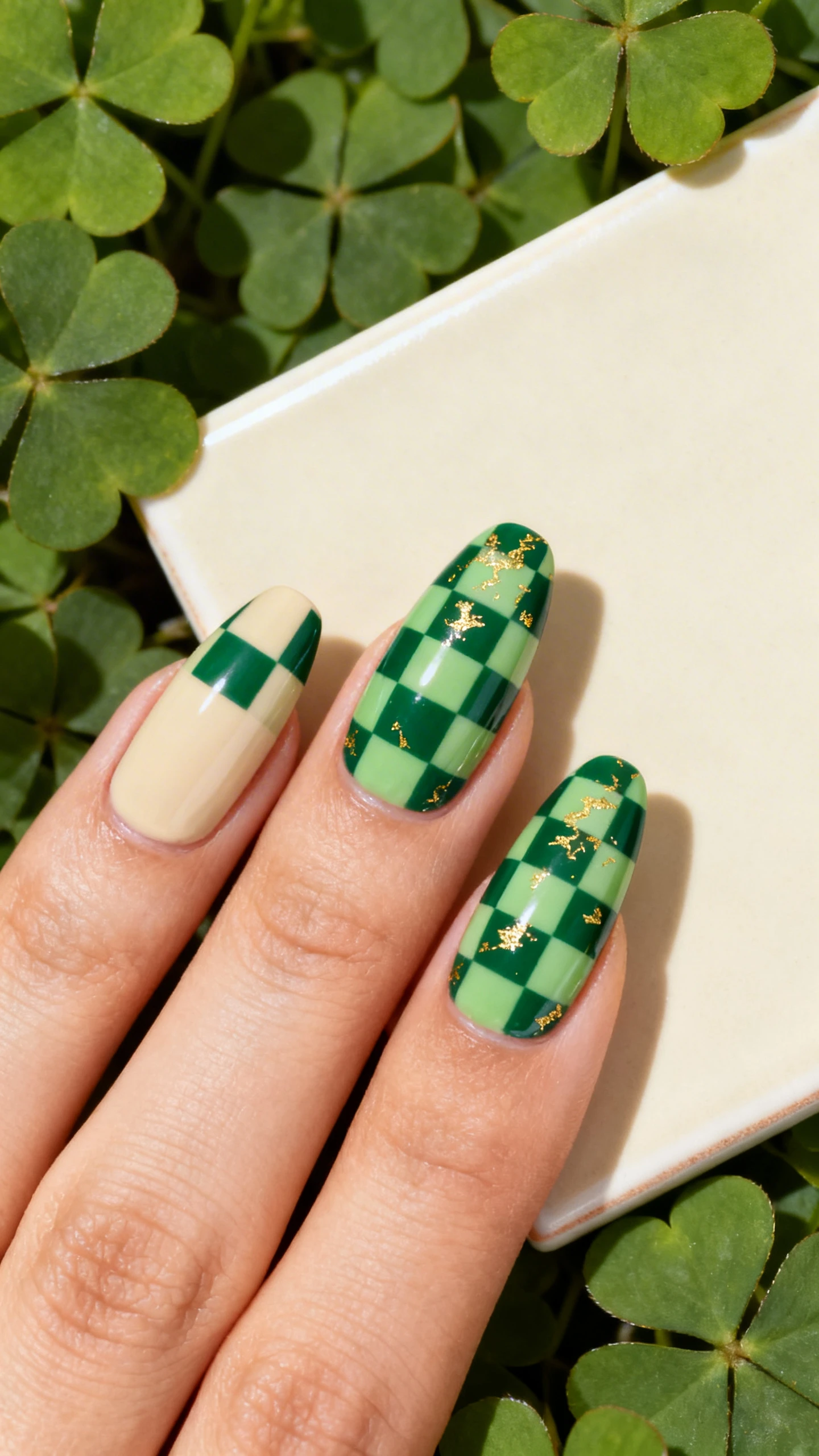 A detailed macro photo of one hand with medium almond nails over an opaque neutral cream base, featuring a bold two-tone checkerboard pattern of forest green and sage green (2x2 squares per nail), topped with a sheer gold flake veil lightly dispersed so the checks remain crisp; razor-sharp focus on the nail art, no faces; background of clover leaves and a matte cream tile for contrast, modern studio lighting with subtle shadow play.
