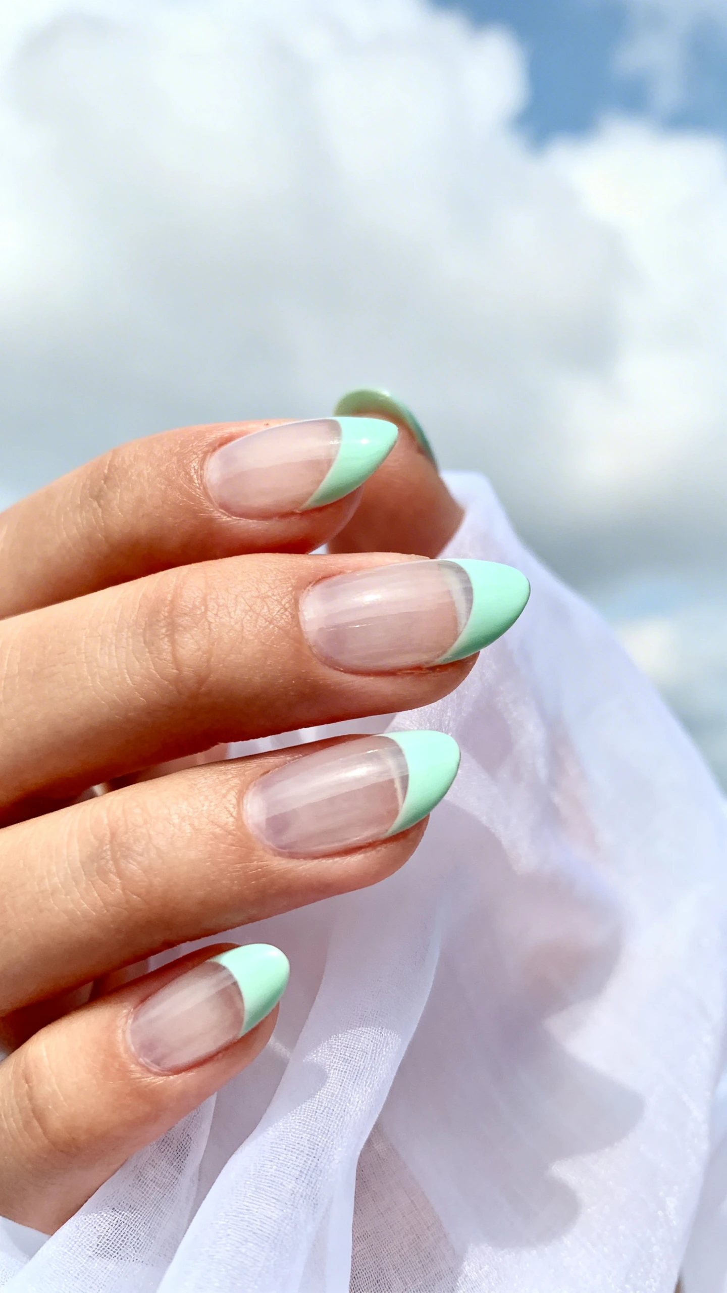 A high-resolution macro photo of one hand with short to medium oval nails, transparent base, and ultra-thin soft mint green French tips 1–2 mm thick; tips follow the natural curve with a slightly blurred, soft-focus edge. Lighting is bright and diffused for crisp detail. Background: airy, pale-cloud backdrop or soft white chiffon evoking a sky-like feel. Modern, Instagram-worthy composition with sharp focus on the nail edges and tip blur. No faces, only the hand, clean cuticles, sheer clear base coat and cool, pastel mint lacquer for tips.