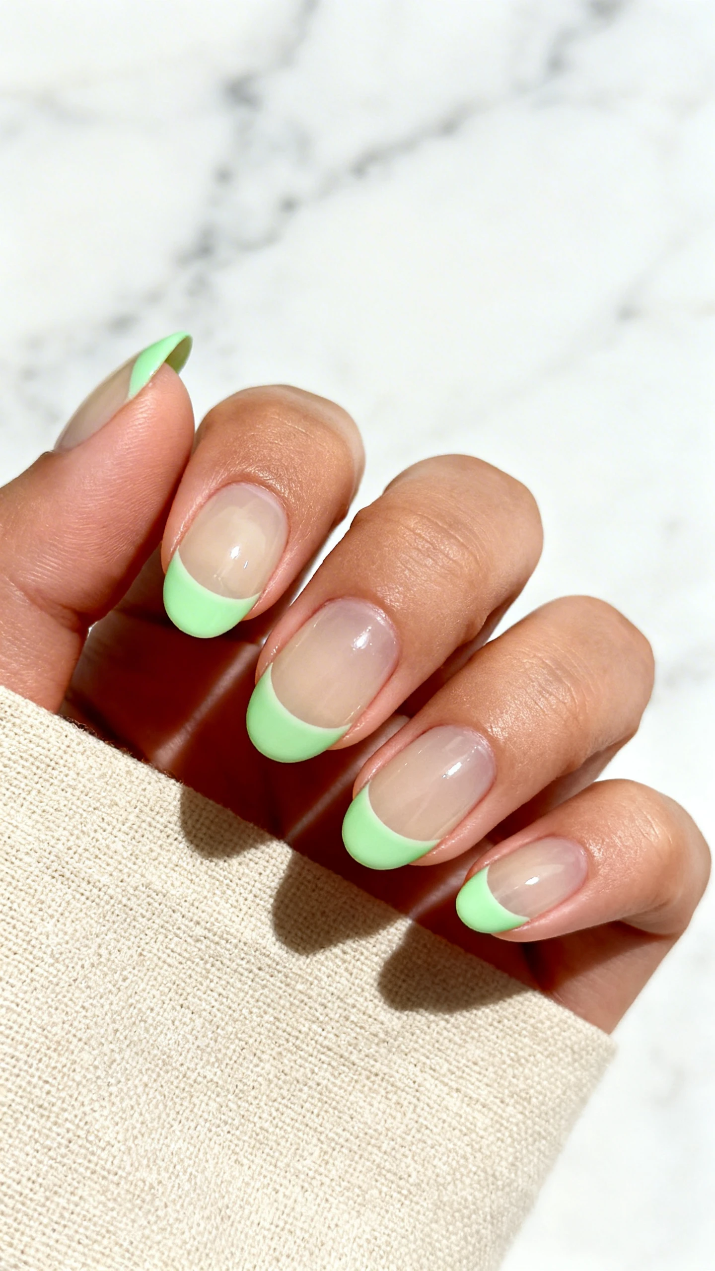 A high-resolution macro photo of one hand with short, softly rounded nails featuring a clear to sheer beige base and precise mint green half-moons 3–4 mm at the cuticle, following the natural curve; rest of each nail remains sheer. Ultra-glossy top coat finish, no glitter. Background: minimalist marble or beige linen to echo the pistachio theme. Clean, modern lighting with sharp focus on the cuticle arcs. Instagram-ready composition, no faces, only the hand.