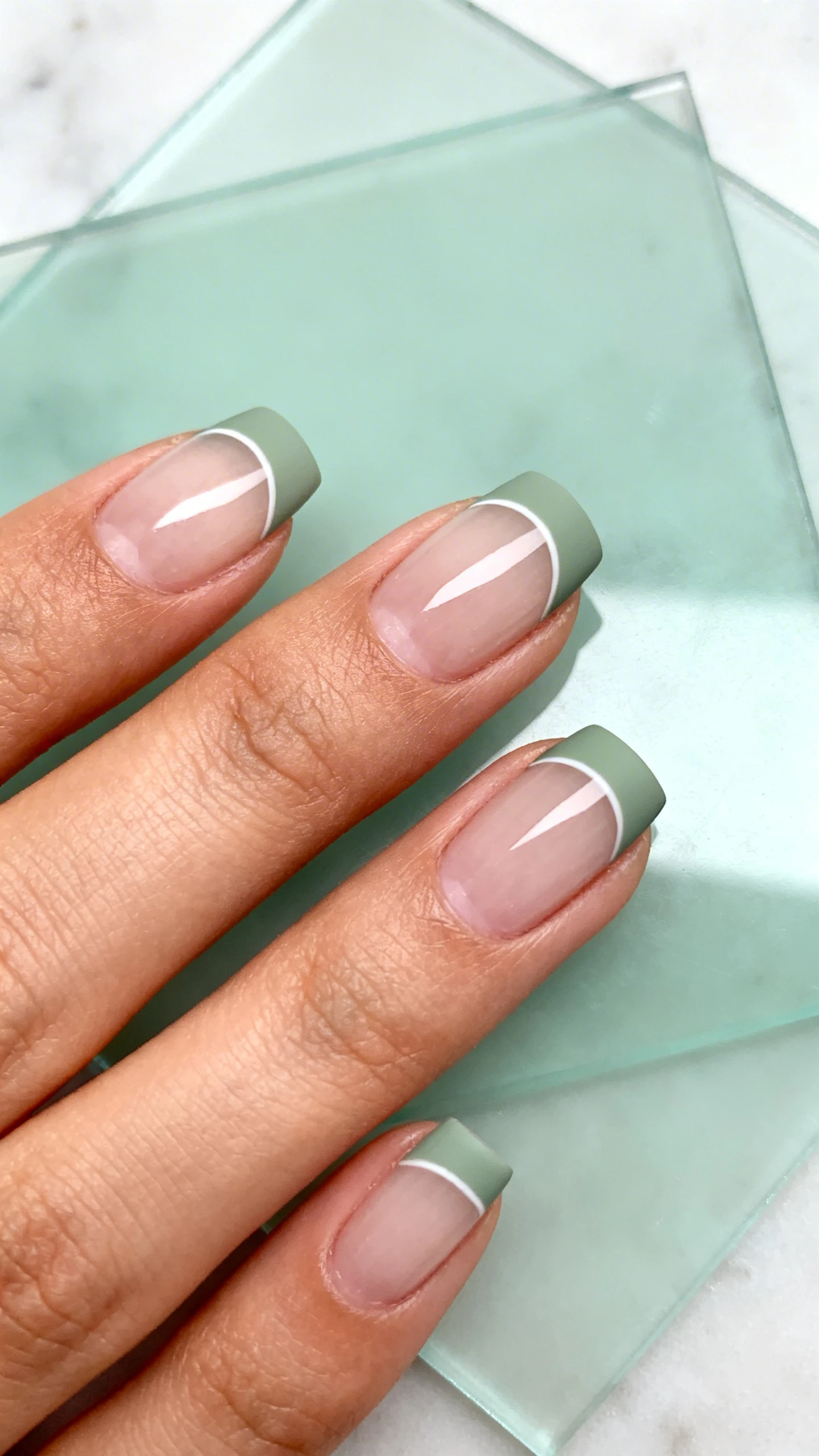 A high-resolution macro photo of one hand with short to medium almond or short square nails, featuring a clear strengthening base only for clean negative space. Add a matte sage French arc on each tip, medium-thin (about 2–3 mm), following the natural smile line with precise symmetry. Highlight the contrast between natural nail and soft matte sage tips. Background: minimal, airy setting like a translucent sage-tinted acrylic sheet or pale marble. Soft, modern lighting; no faces, only the hand and nails.