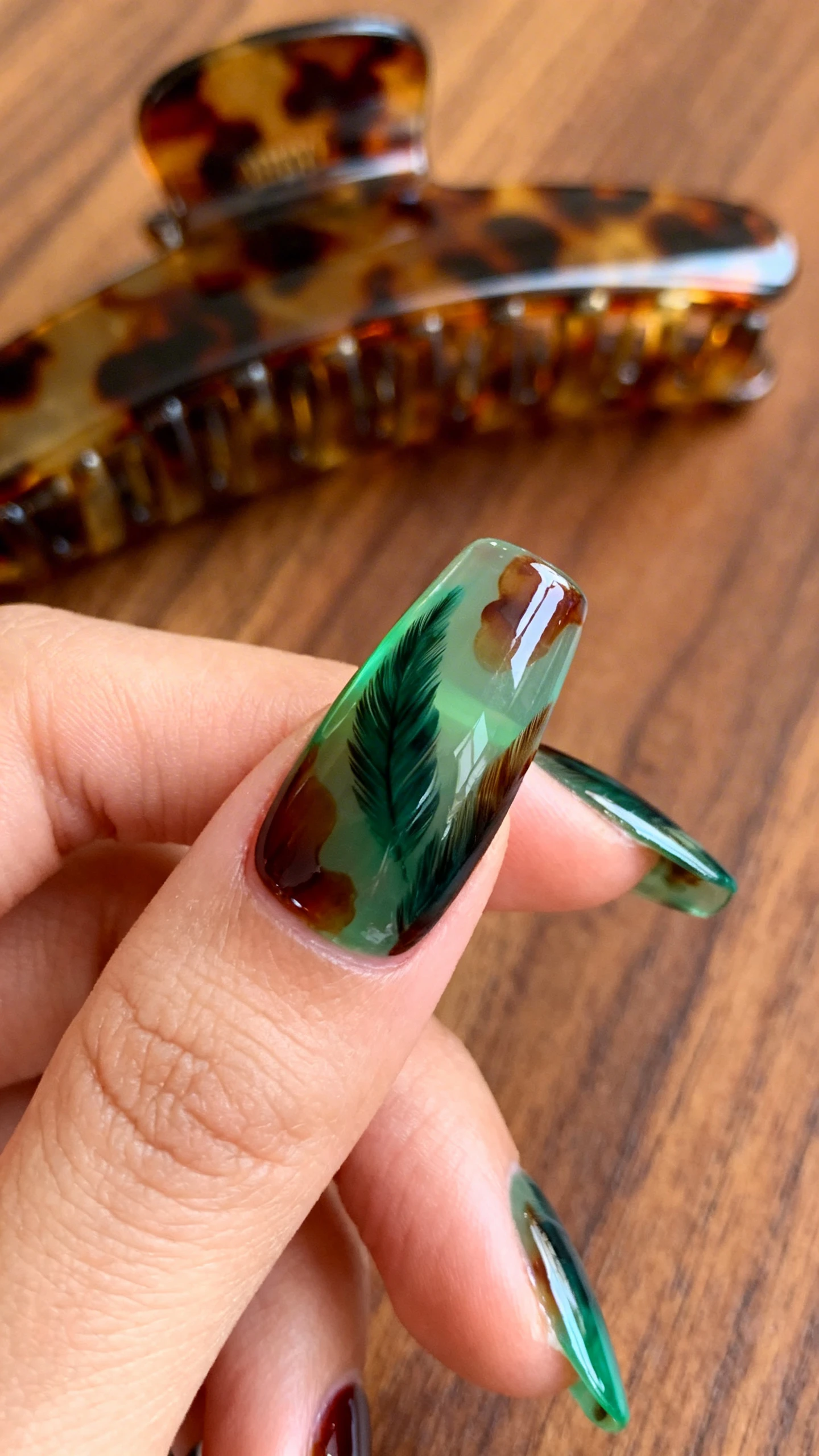 A macro, high-resolution photo of one hand with medium squoval nails featuring an emerald tortoiseshell design: a translucent emerald jelly base layered with irregular, feathered swirls of dark forest green and cola-brown gel, softly overlapped to create depth; glossy top coat for a luxe finish; photographed on a warm walnut wood surface with a vintage tortoiseshell hair accessory slightly out of focus in the background, modern editorial look, sharp nail detail, no faces.