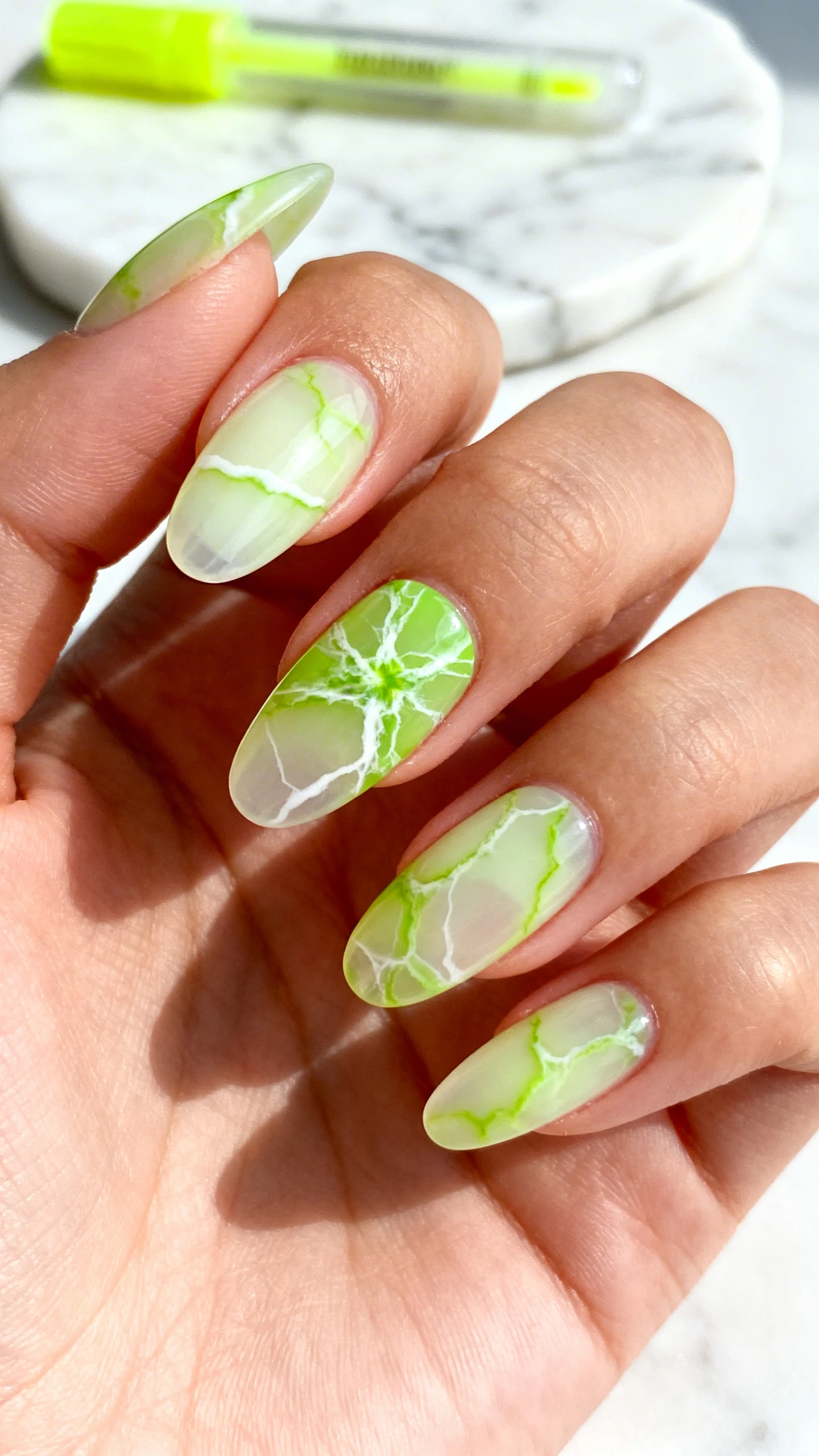 Macro photo of a single hand with medium almond nails in Highlighter Marble Veins: semi-sheer milky cream base; layered wisps of neon green marbling flowing from cuticle to tip; fine soft white veins interlaced to mimic natural stone; veining varies in thickness and is slightly blurred; no glitter. One accent nail with slightly denser veining at the center. Lighting: diffused daylight to emphasize translucency and depth. Background: white marble slab with a neon highlighter pen softly out of focus. No faces; only hand; high-res, crisp focus on marbling texture; clean Instagram look.