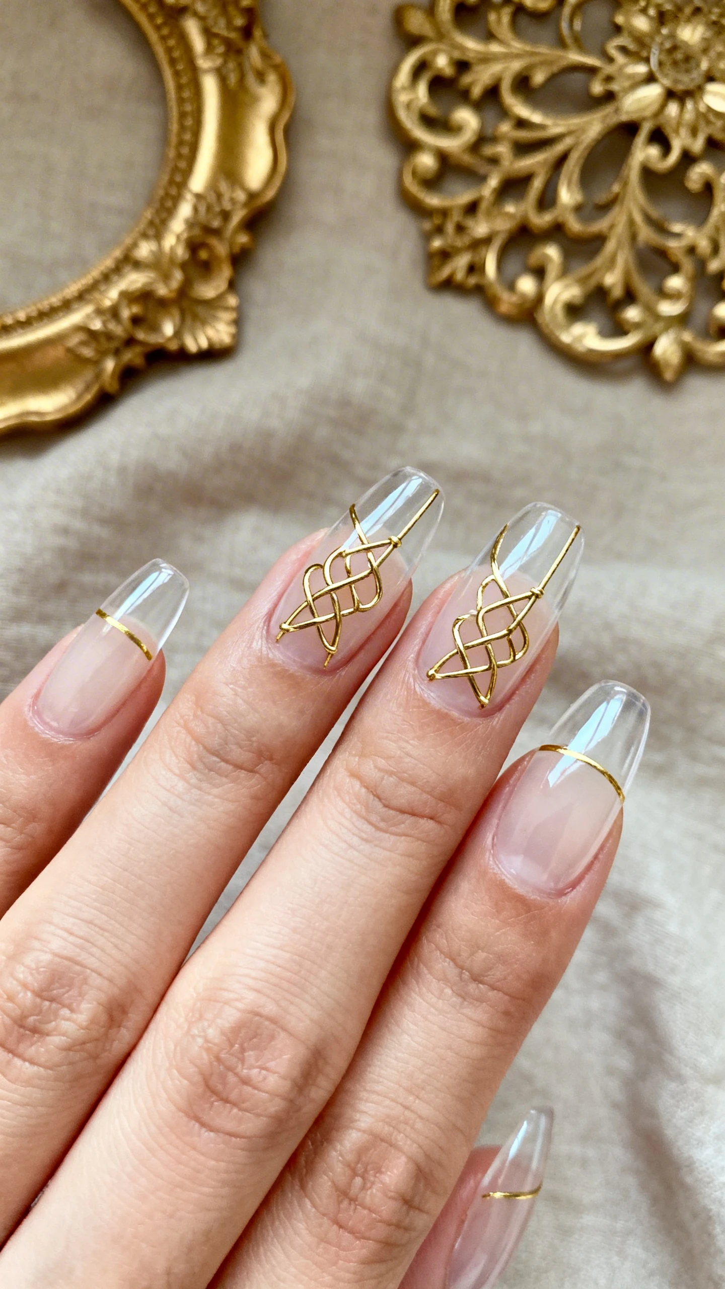 Macro photo of one hand with a transparent, high-gloss clear base (or ultra-sheer cream wash); 2 accent nails (ring and middle) display thin metallic gold interlacing lines forming Celtic knot-inspired patterns—about 1 mm thick, with small looped intersections and soft angles; remaining nails feature ultra-fine gold linear details (subtle arcs or single stripes) for balance; high-resolution, tack-sharp lines; aesthetic background: antique gold filigree jewelry on a soft neutral cloth; chic studio lighting; no faces, only the hand and nails.