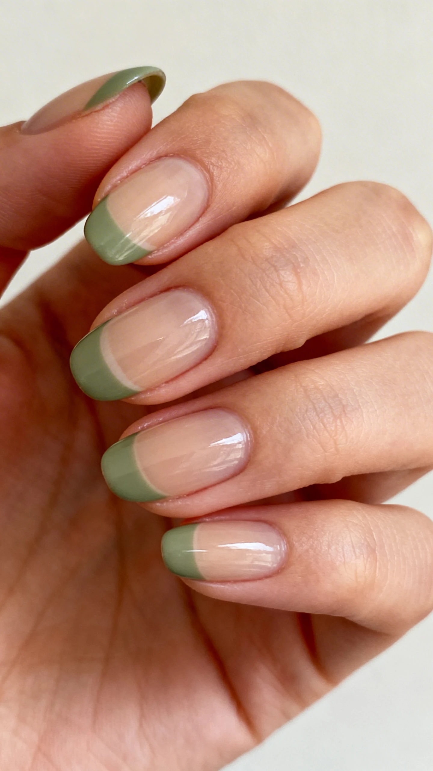 Macro photo of one hand with medium almond nails, sheer neutral base preserving natural warmth. French tips in muted sage green, medium thickness (