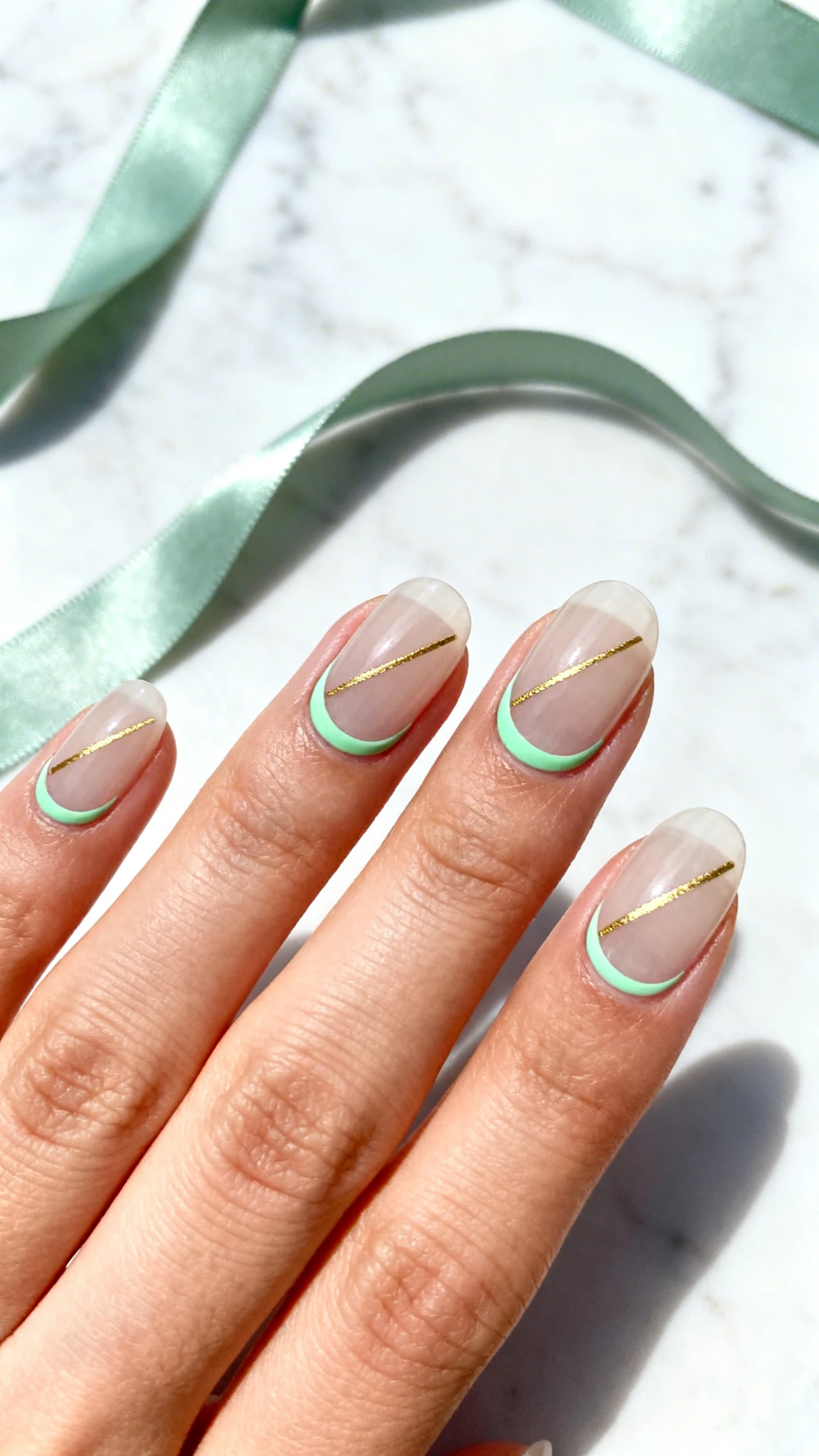 Macro photo of one hand with medium-short oval nails, translucent neutral base wash; delicate crescent moon outlines in soft sage green hugging the cuticle curve (approx. 2–3 mm thick); each nail features one ultra-fine metallic gold diagonal “lucky line” running from lower sidewall toward the free edge; minimal, elegant negative space; high-resolution, crisp focus; background: pale marble with a faint sage silk ribbon for a refined Irish nod; bright, natural light aesthetic; no faces, composition centered on the cuticles and gold lines.