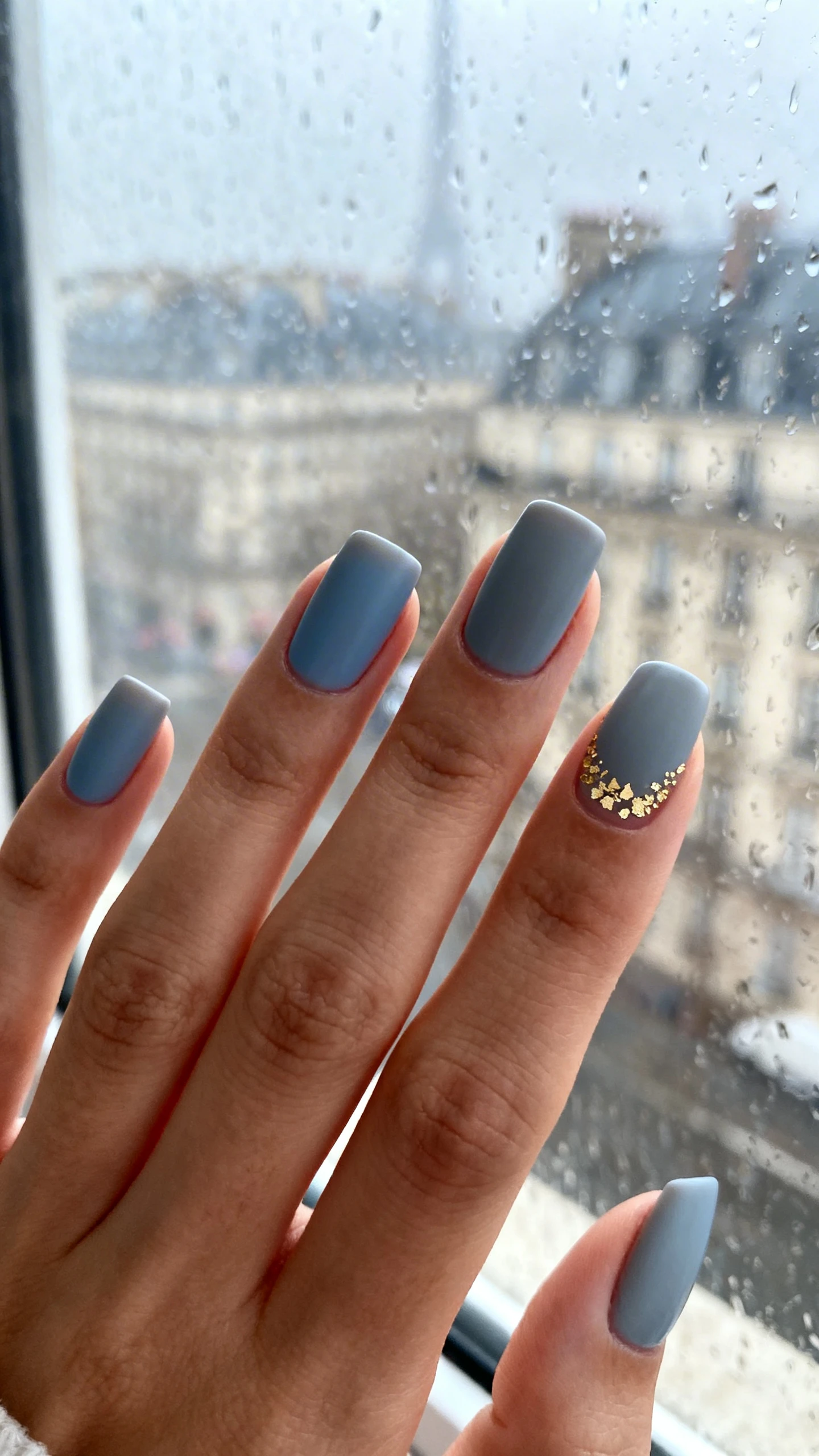 Macro photo of one hand with medium-short square nails with softened corners, painted opaque cool gray with a soft blue undertone (Paris fog); index, middle, and pinky are solid gray, no embellishment; ring finger and thumb feature the same gray base with a delicate veil of fine, irregular gold foil specks clustered in a crescent near the cuticle fading outward, ultra-refined and not chunky; high-resolution, sharp focus to capture foil texture and smooth polish; background: rainy Paris window vibe—cool-toned blurred cityscape or misty glass; sleek editorial lighting; no faces, only the hand and nails.
