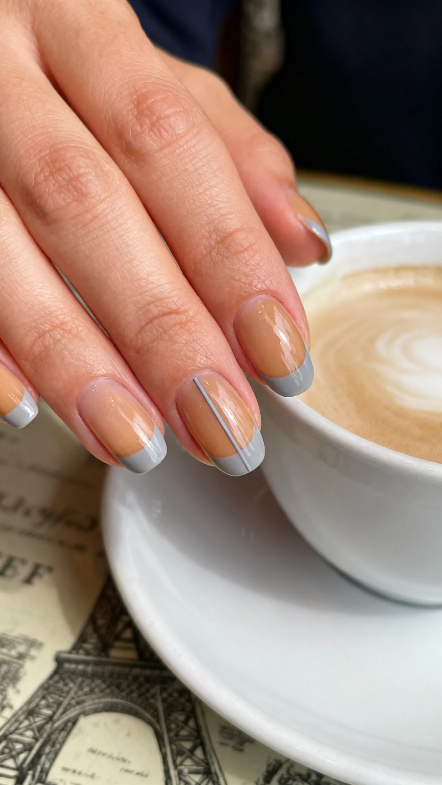 Macro photo of one hand with short to medium oval nails, sheer café-crème base with a warm café au lait translucence; tips feature a crisp 1–2 mm micro-French in pale dove gray perfectly following the natural curve; ring finger shows an ultra-fine, perfectly straight vertical dove-gray line centered from cuticle to free edge; high-resolution, sharp focus capturing the gloss and micro-French precision; background: softly blurred Parisian café tabletop with a porcelain cup and saucer in neutral tones; modern, Instagram-worthy lighting with gentle daylight; no faces, only the hand and nails.