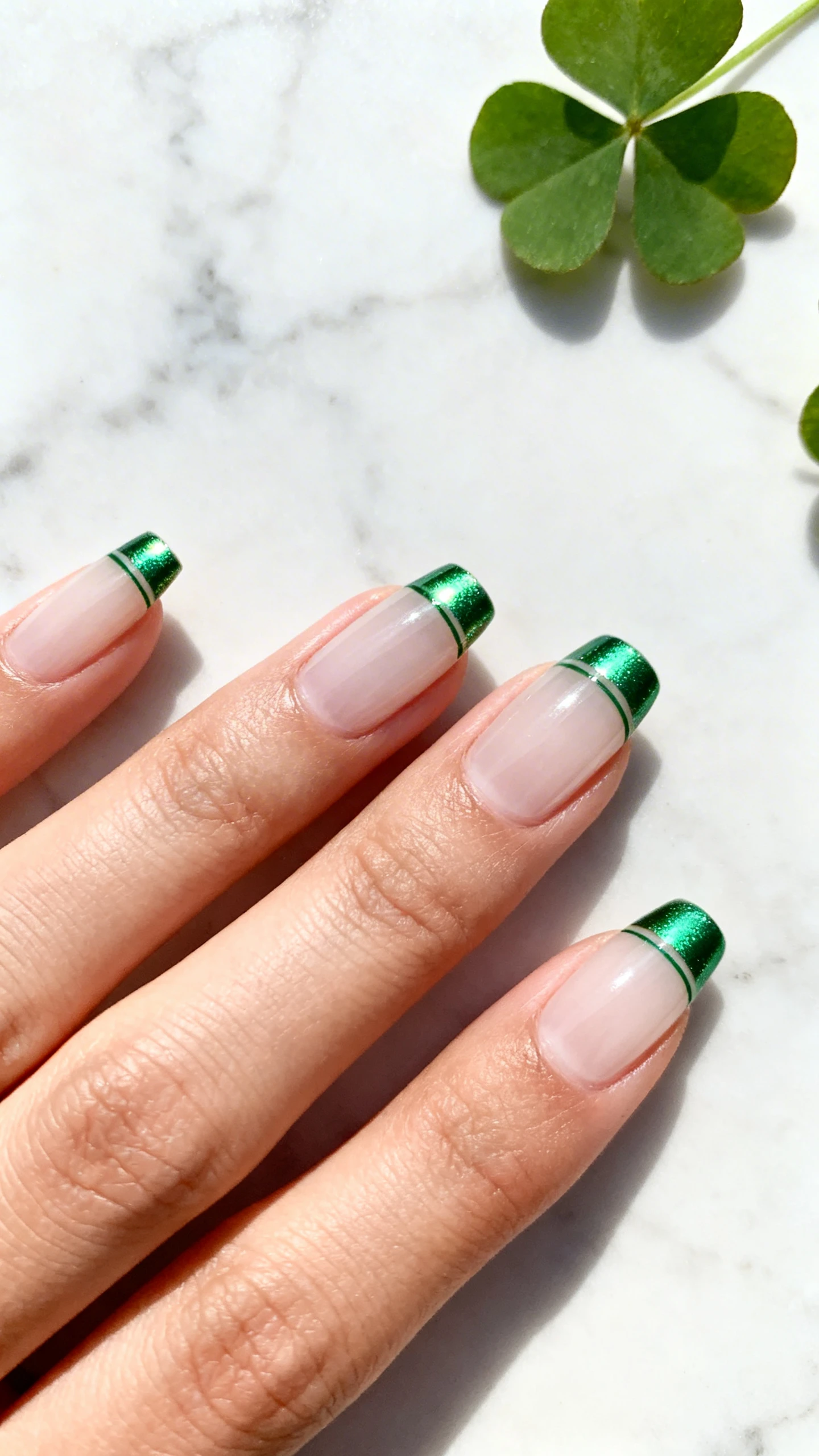 Macro photo of one hand with short-to-medium rounded/soft square nails featuring a sheer milky neutral base (semi-translucent, no pink) and ultra-thin metallic shamrock green micro-French tips with a reflective foil effect; razor-sharp focus on the delicate tip line and clean sidewalls; background of pale marble and a hint of clover leaves for a chic St. Patrick’s theme; bright, natural studio lighting with minimal shadows; no faces, only the hand and nails.